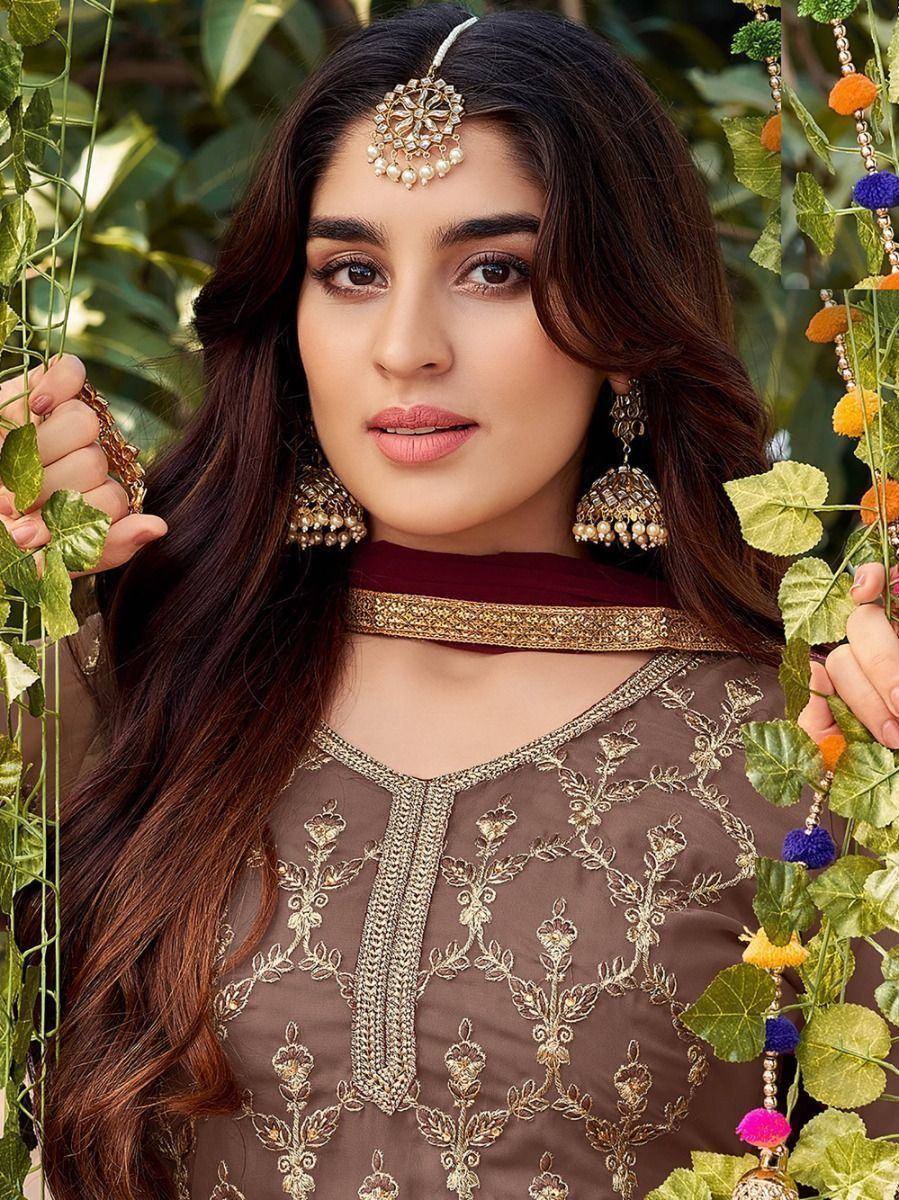 Woman in embroidered brown ethnic gown with gold jewelry, against leafy outdoor backdrop
