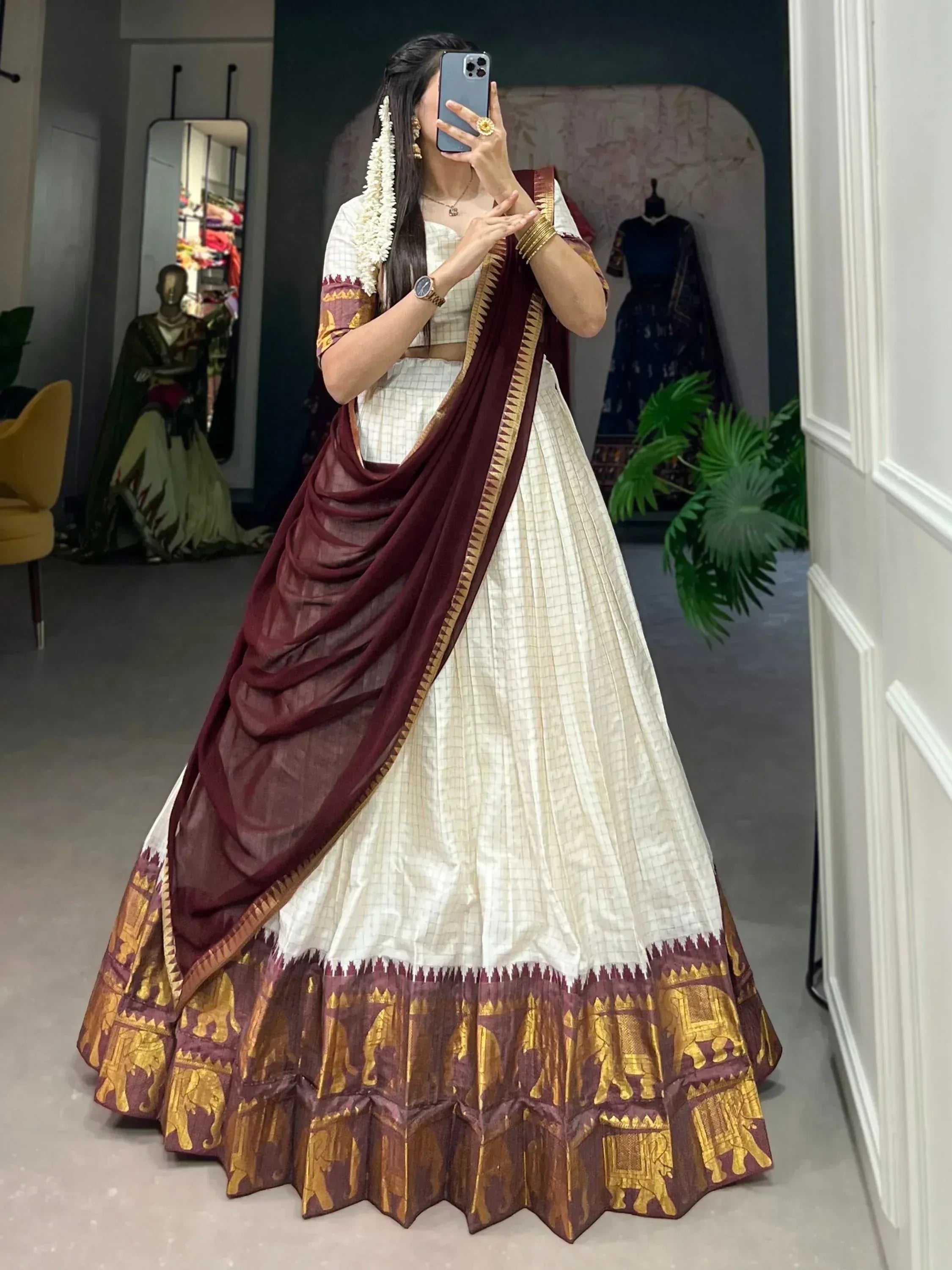 Woman in white and maroon lehenga choli with gold elephant print, taking mirror selfie in boutique