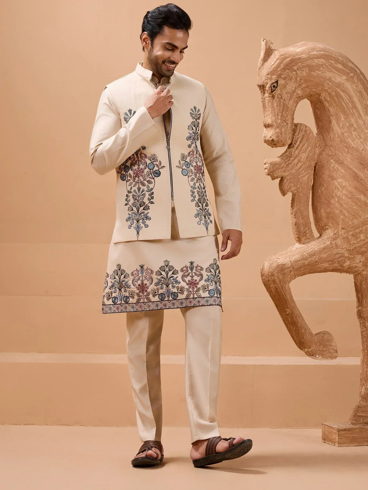 Man in cream embroidered kurta set with floral motifs, paired with matching pants and sandals, beside a wooden horse sculpture.