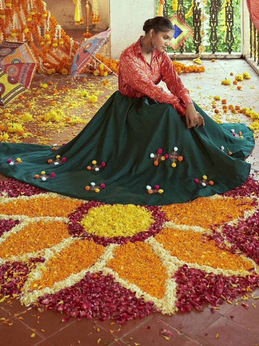 Woman in red printed top and green embroidered skirt seated on floral rangoli, festive Indian ethnic wear