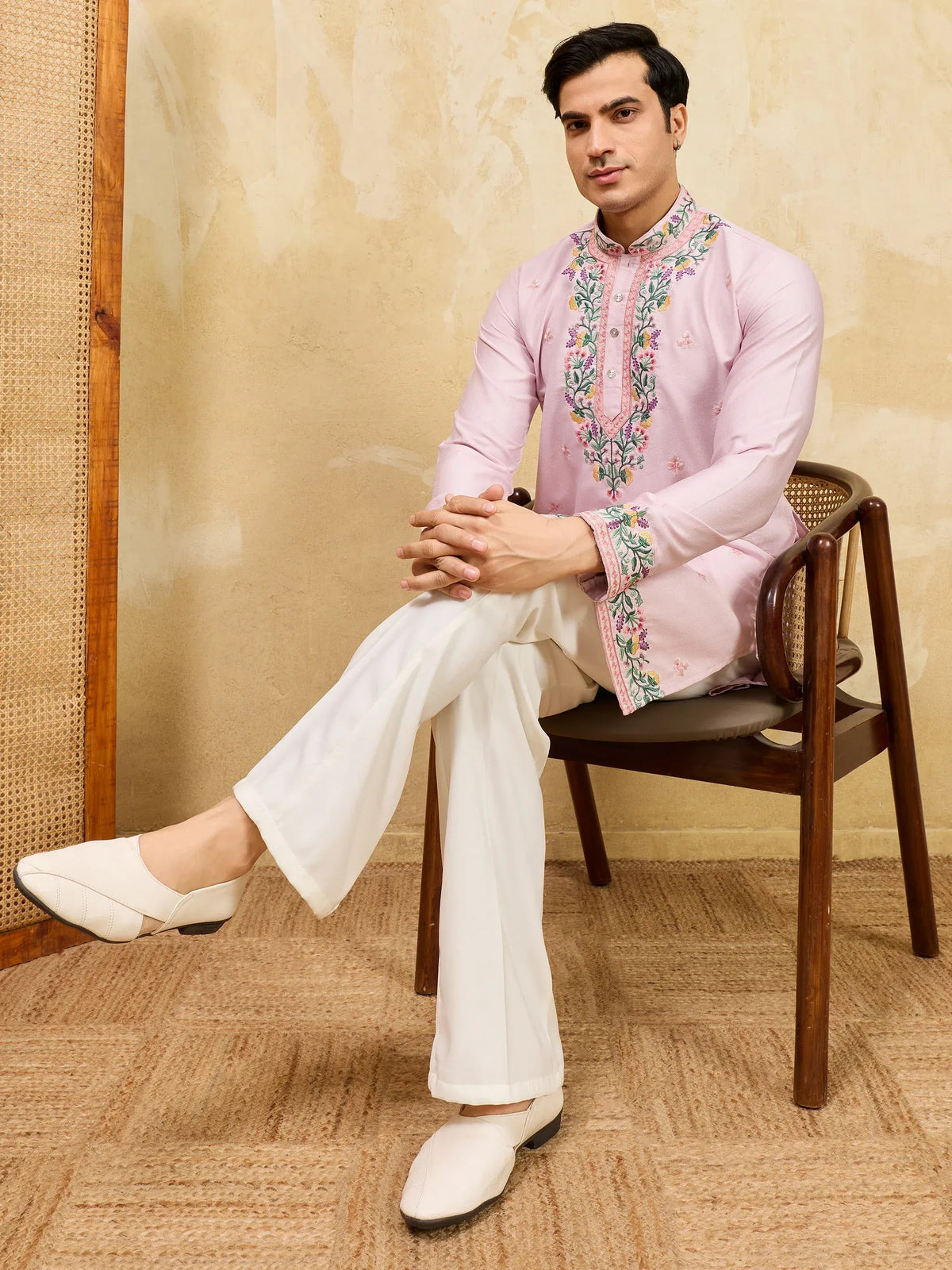 Man in a pink embroidered ethnic kurta and white trousers sitting on a wooden chair.