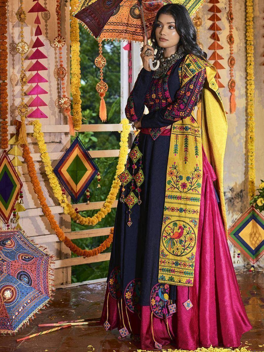 Woman in embroidered navy and magenta ethnic gown with yellow dupatta, holding decorative umbrella, festive Indian decor