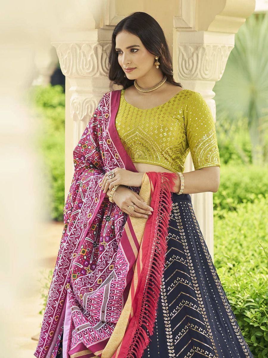 Woman in vibrant Indian lehenga choli with embroidered green blouse, patterned pink dupatta, and navy skirt, styled for ethnic occasions.