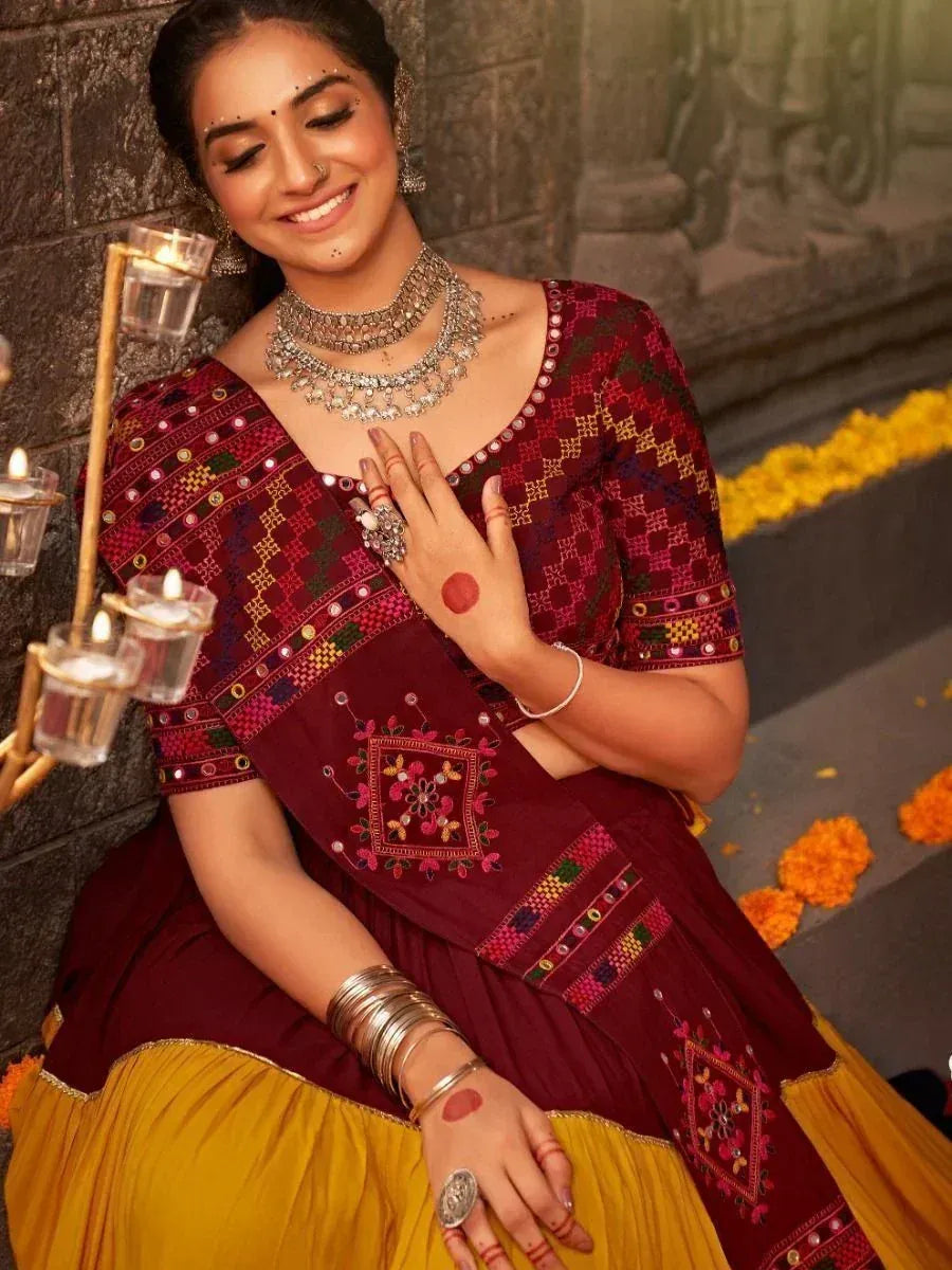 Woman in maroon and yellow embroidered ethnic gown with mirror work, traditional jewelry, and henna hands