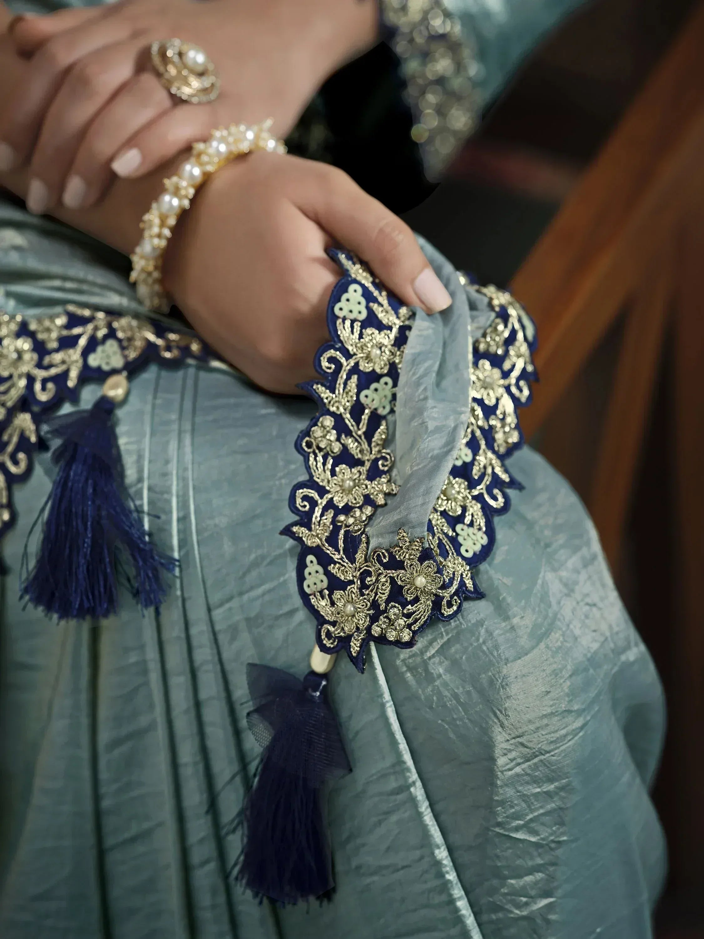 Elegant Indian ethnic wear with gold embroidery, pearl details, and blue tassels on a teal gown.
