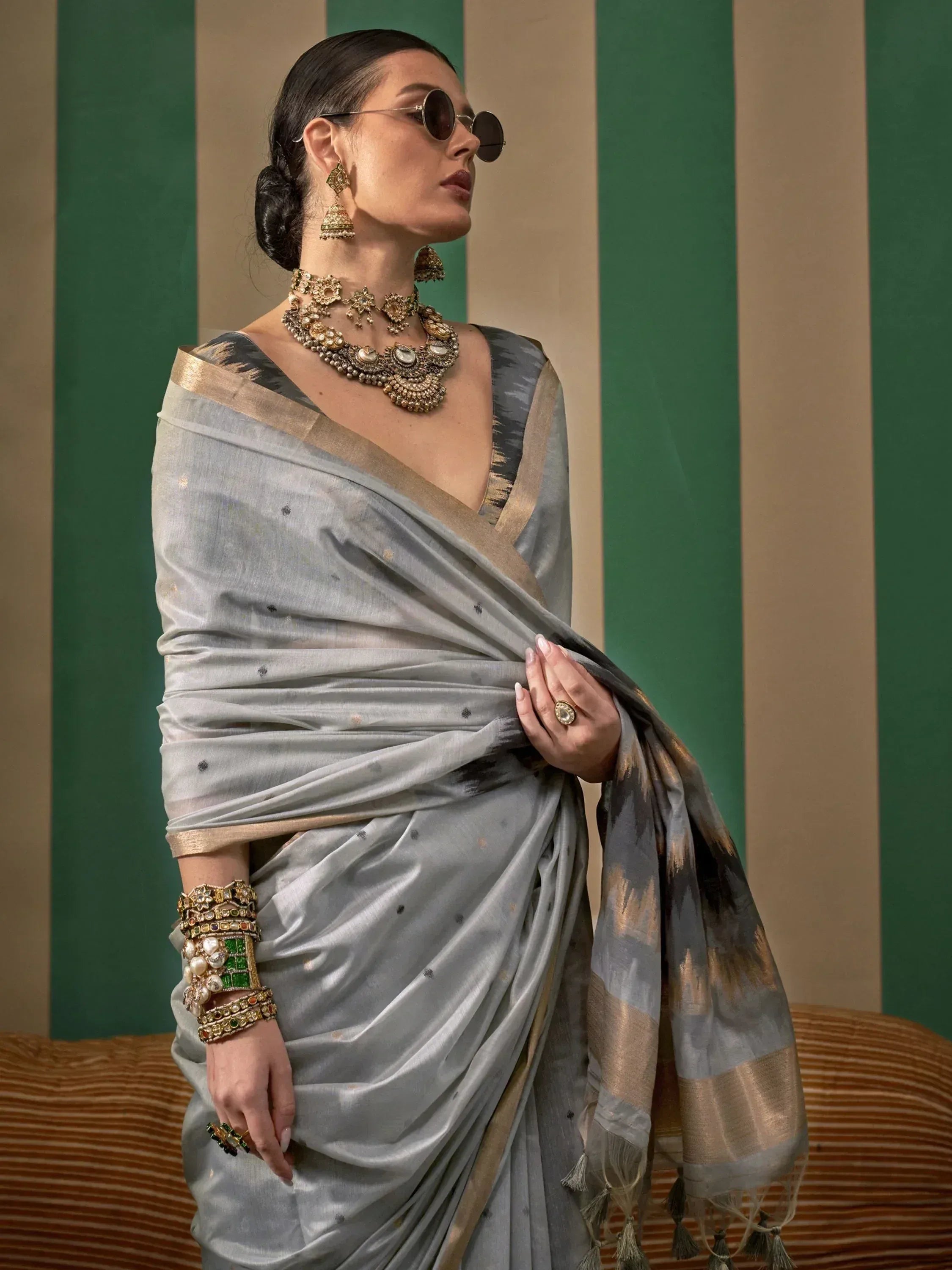 Stylish woman in grey silk saree with gold border, traditional Indian jewelry, and sunglasses against striped backdrop