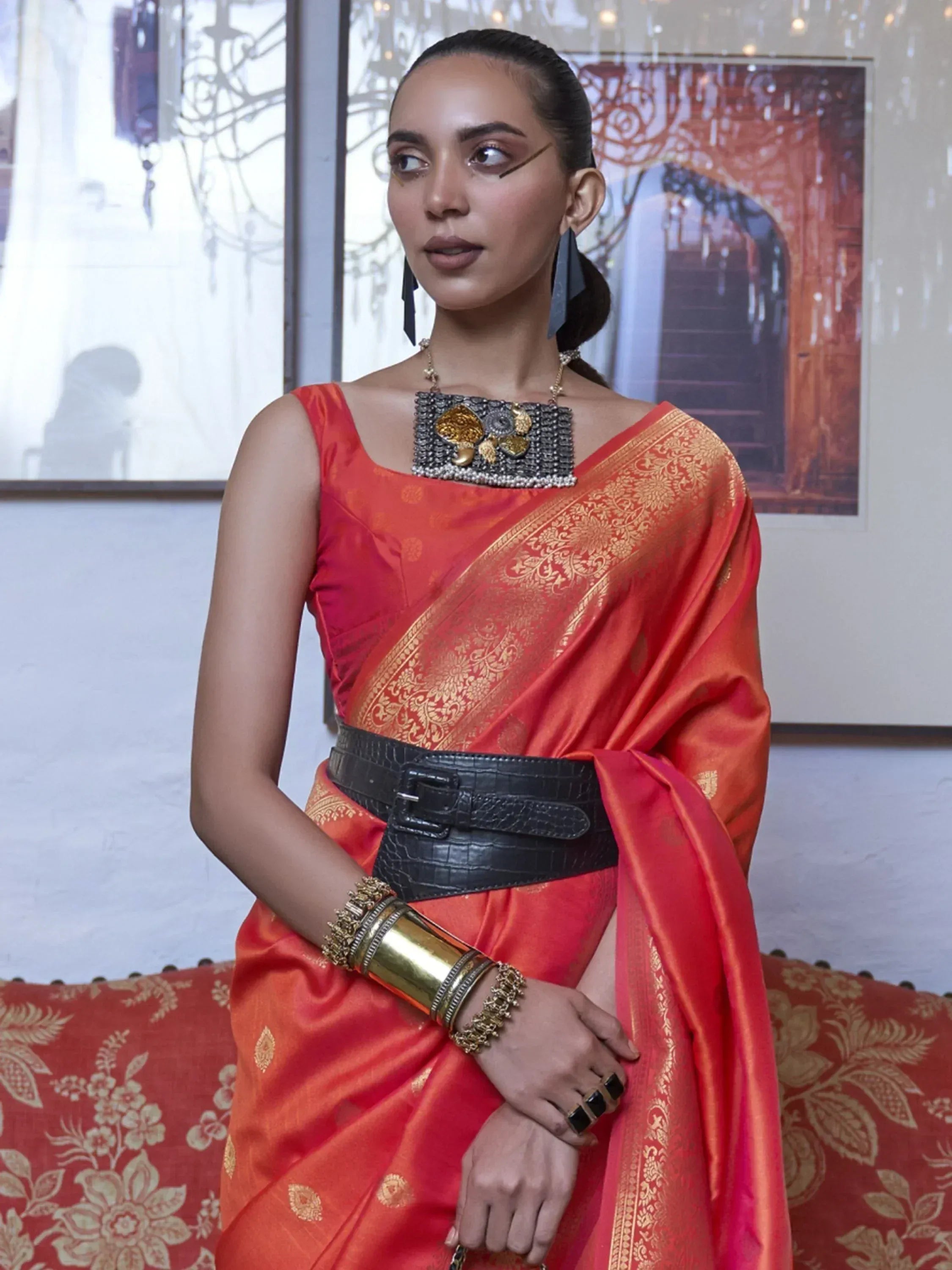 Woman in vibrant orange silk saree with gold embroidery, styled with black belt and statement jewelry for ethnic wear