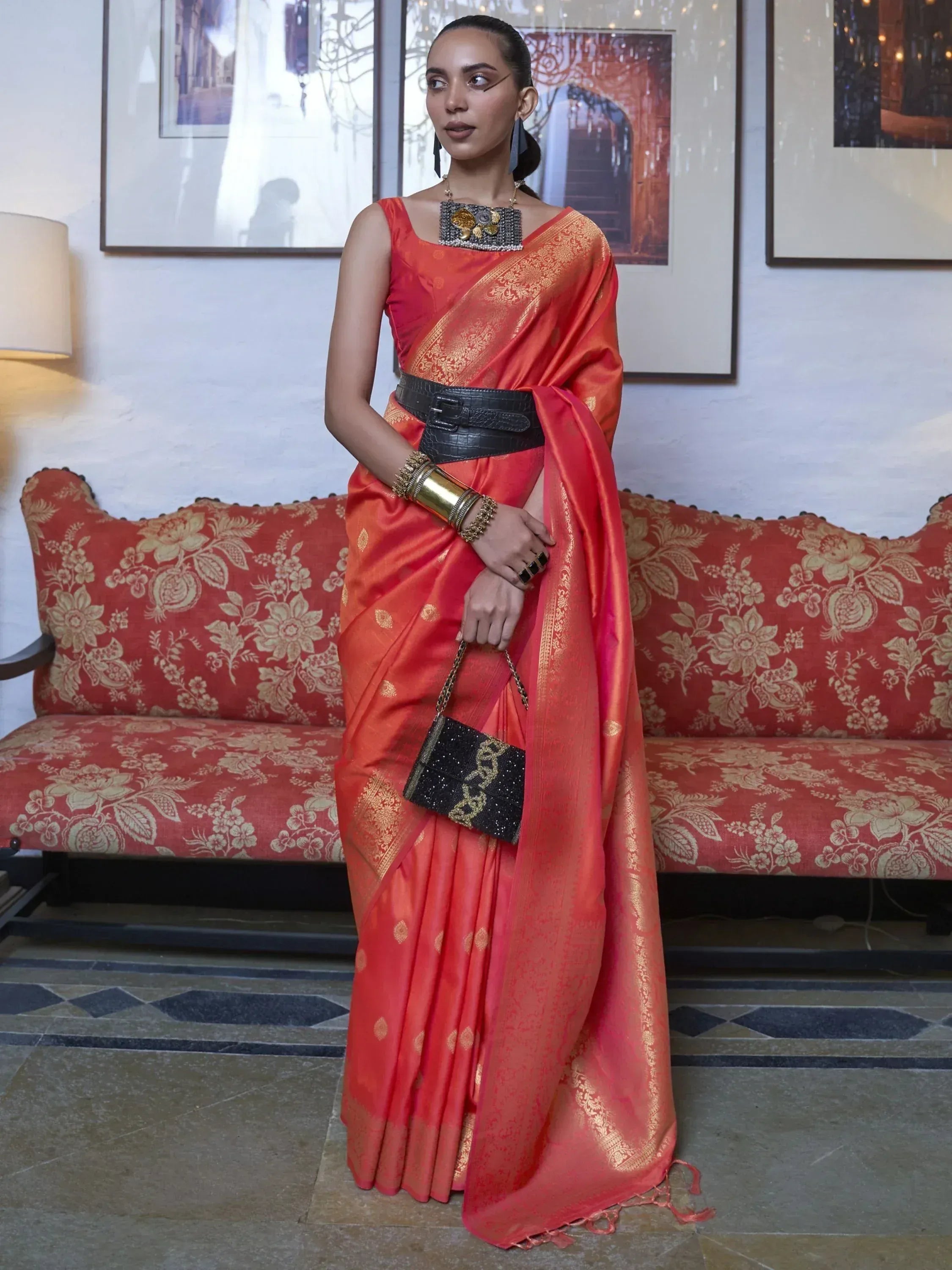 Woman in vibrant orange silk saree with gold zari, black belt, and statement jewelry, styled for ethnic occasions.
