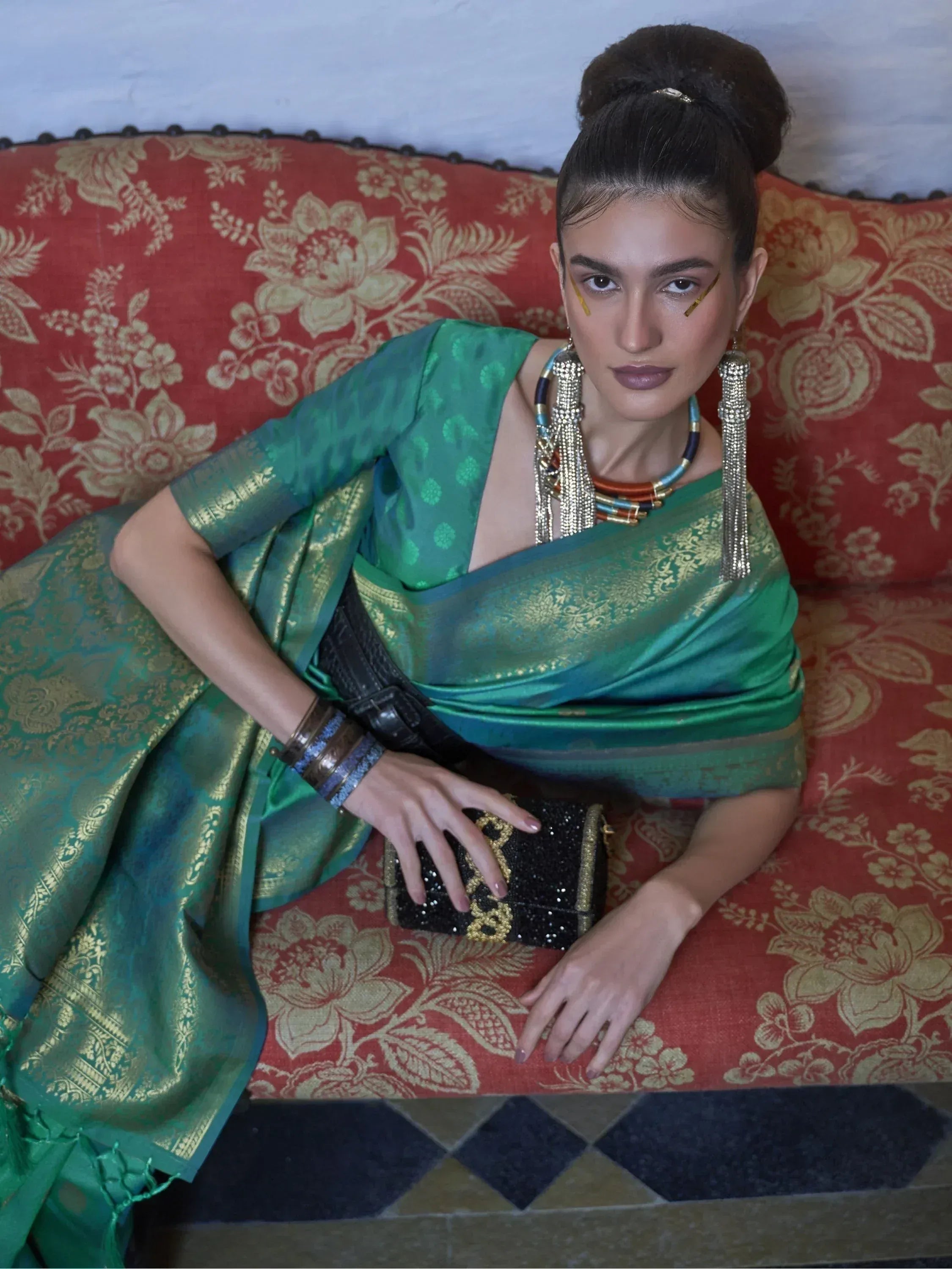 Woman in elegant green designer saree with gold embroidery, styled with statement jewelry and clutch, seated on floral sofa.
