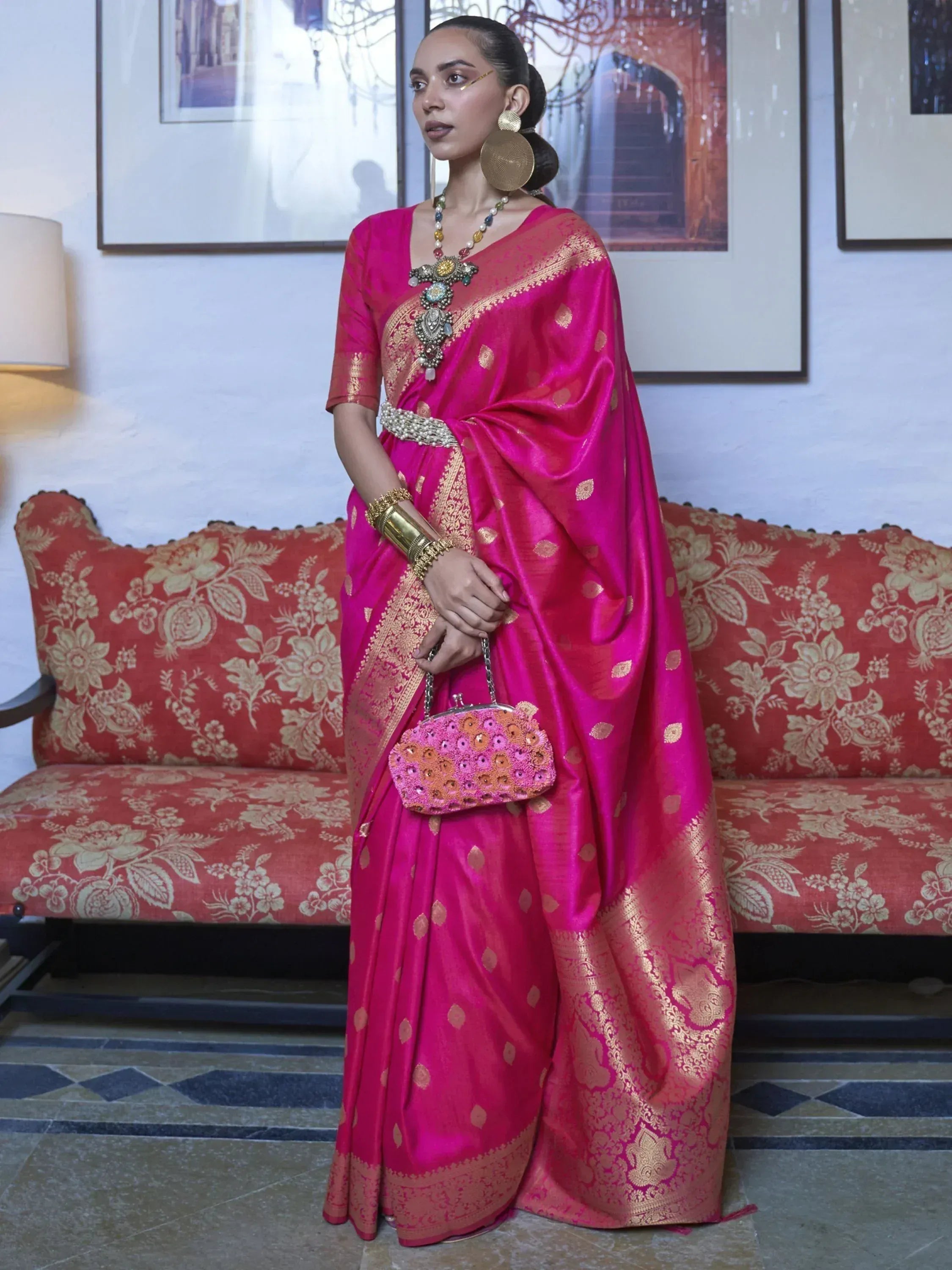 Woman in vibrant pink silk saree with gold zari, traditional jewelry, and embroidered clutch, styled for ethnic occasions.