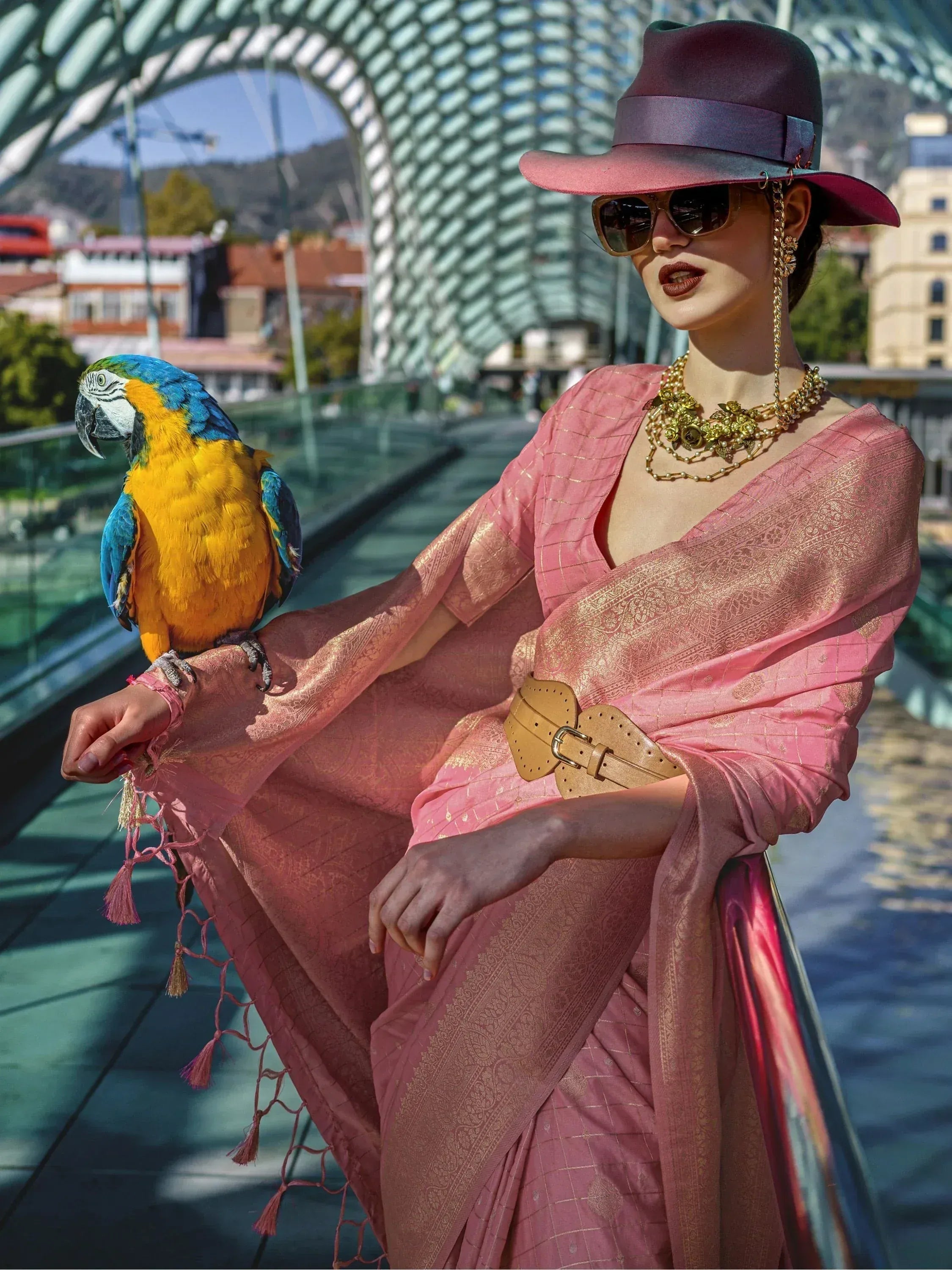 Woman in pink ethnic saree with gold detailing, wearing hat and sunglasses, parrot perched on arm
