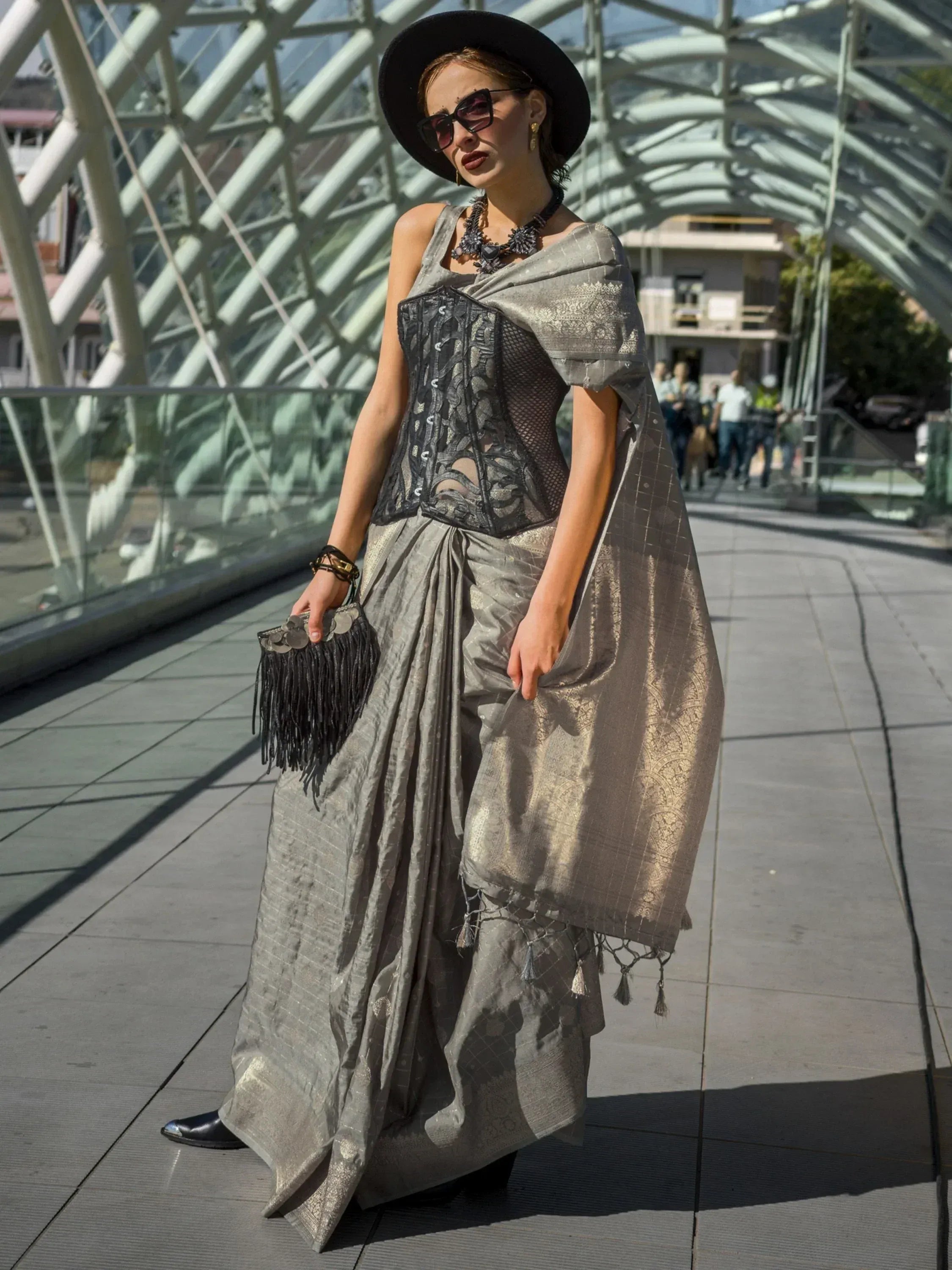 Stylish woman in silver ethnic gown with black corset, wide-brim hat, and fringe clutch on modern bridge