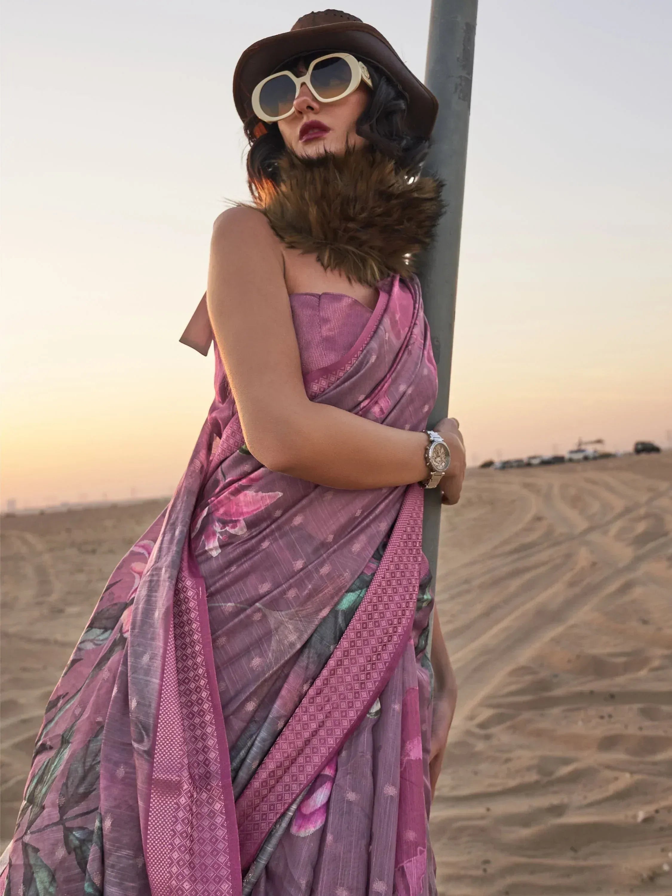 Woman in purple floral saree with fur stole, hat, and sunglasses at desert sunset