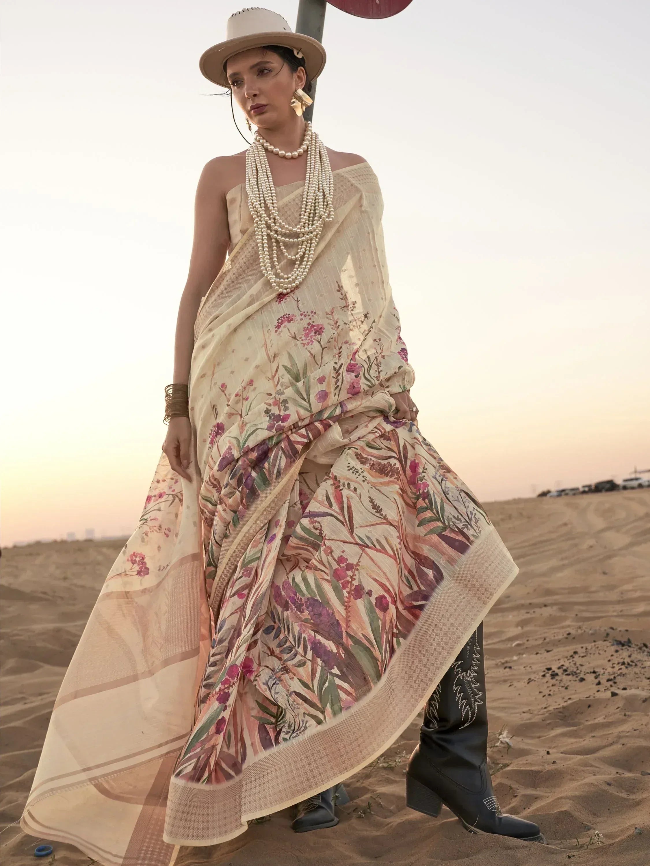 Floral print saree with off-shoulder drape, pearl necklace, and cowboy boots on desert sand at sunset