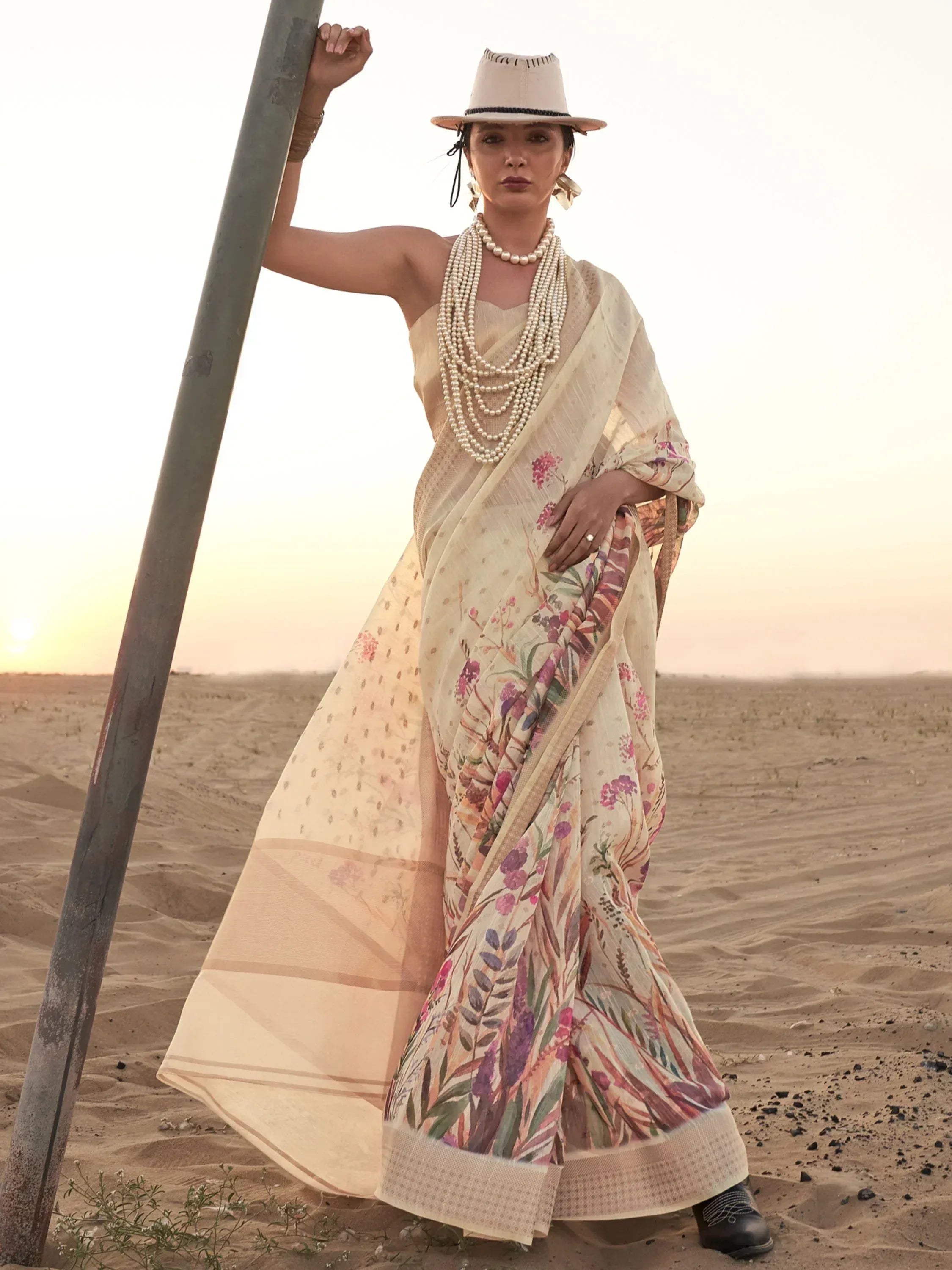 Woman in floral print saree with pearl necklace, hat, and boots in desert at sunset