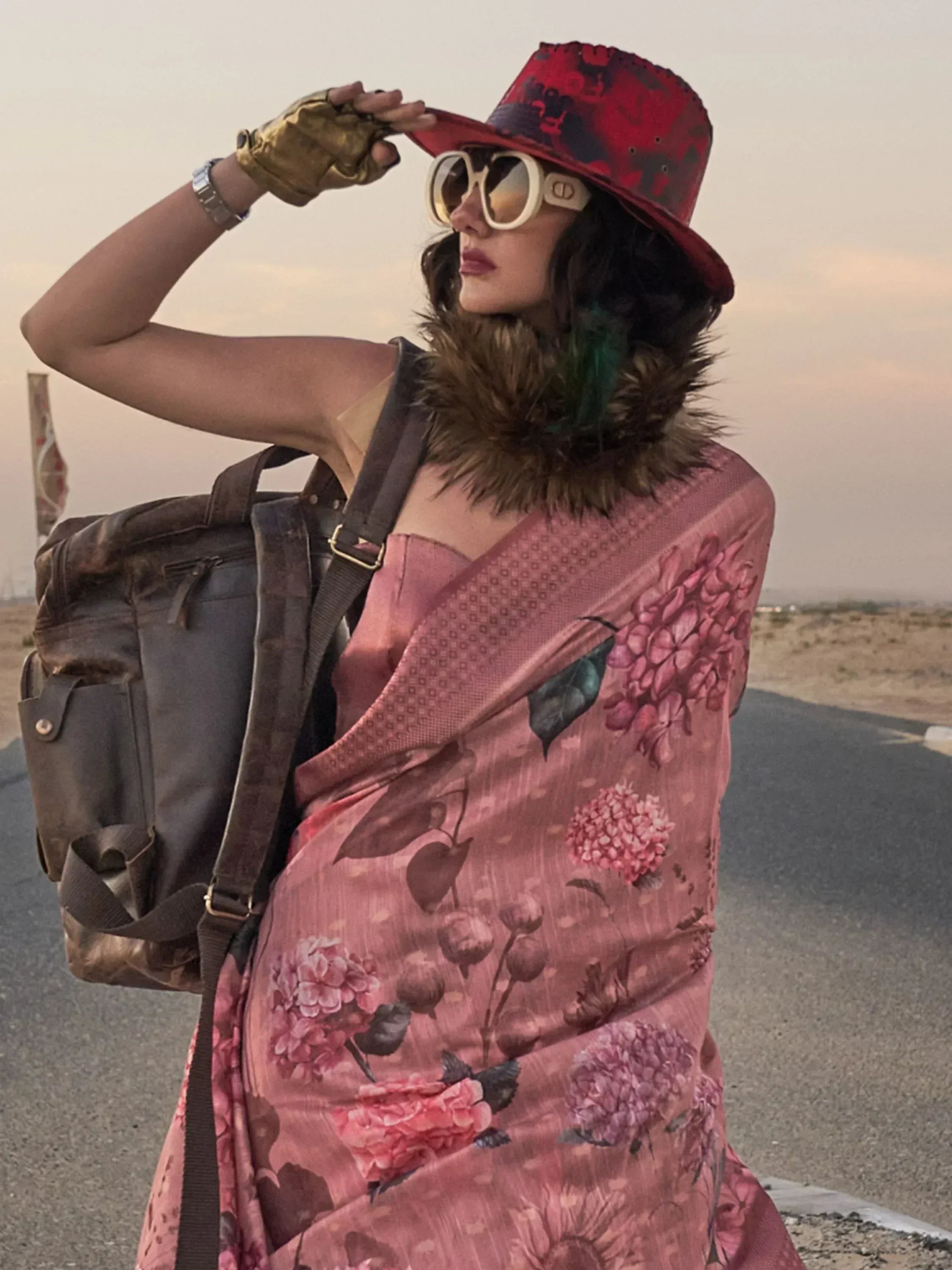 Woman in pink floral saree with red hat, sunglasses, and fur collar, posing on desert road