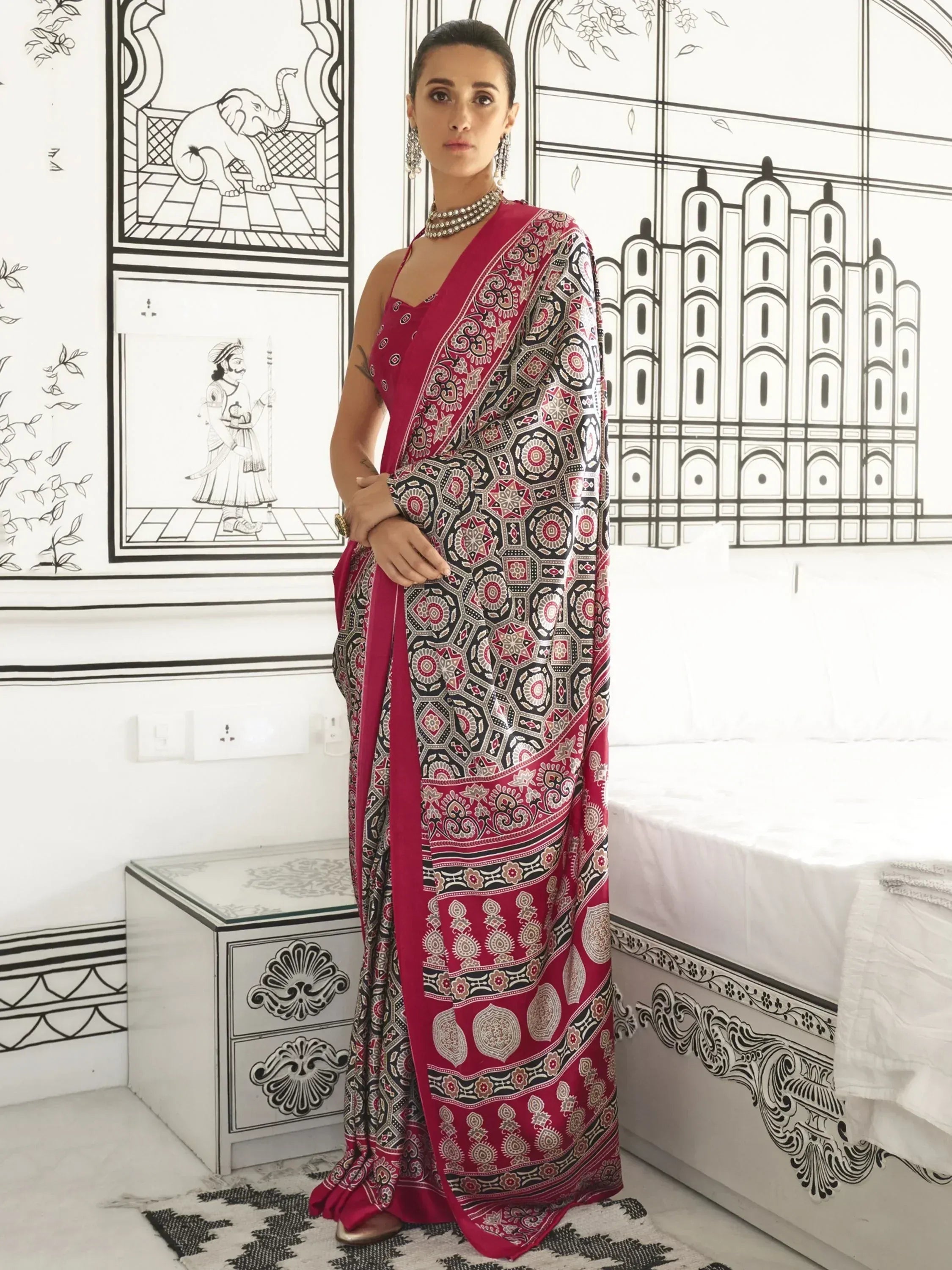 Woman in red and black printed saree with ethnic motifs, styled with pearl jewelry against illustrated Indian architecture backdrop