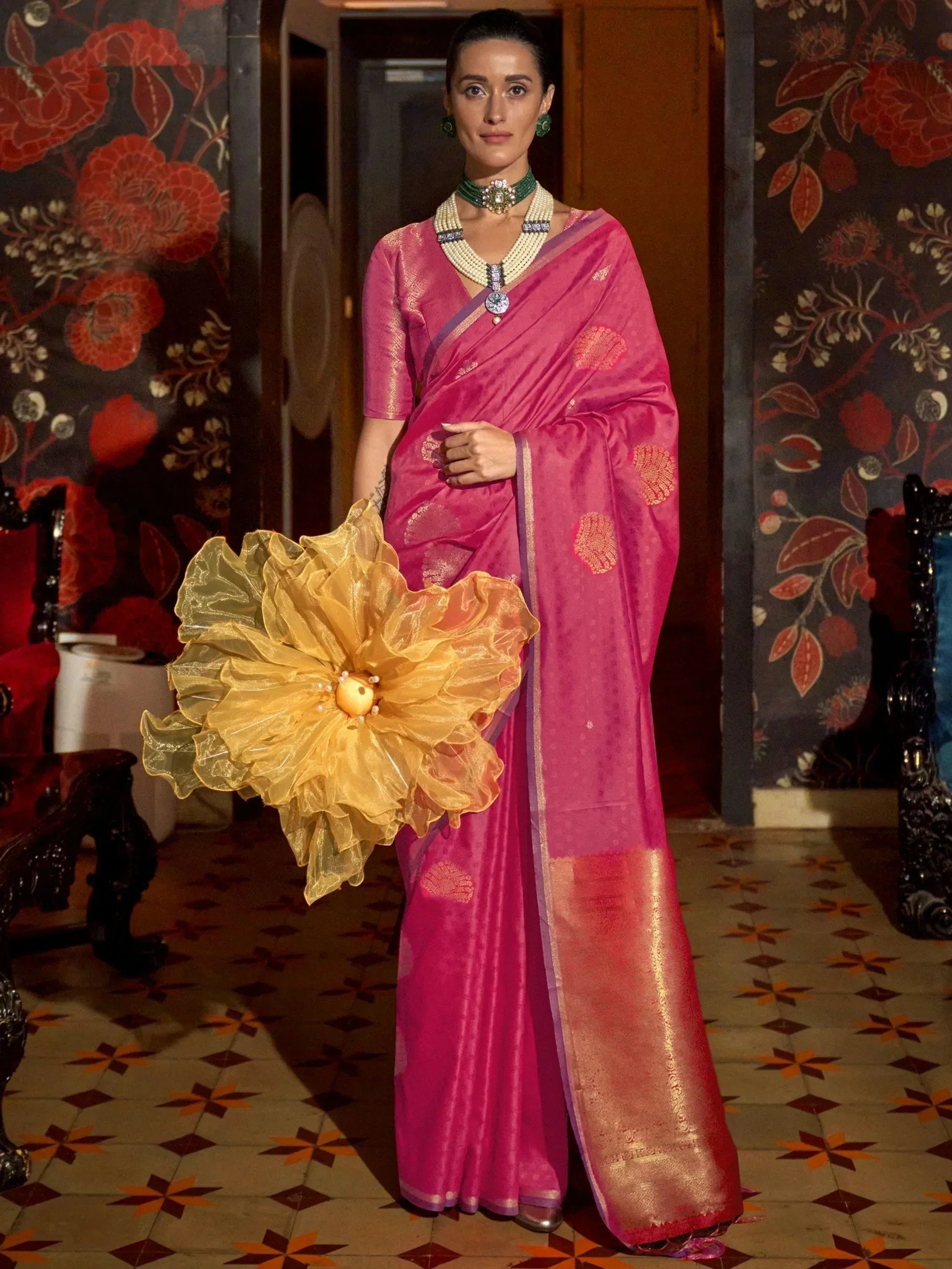 Woman in pink designer saree with gold motifs, holding yellow floral accessory, traditional Indian ethnic wear