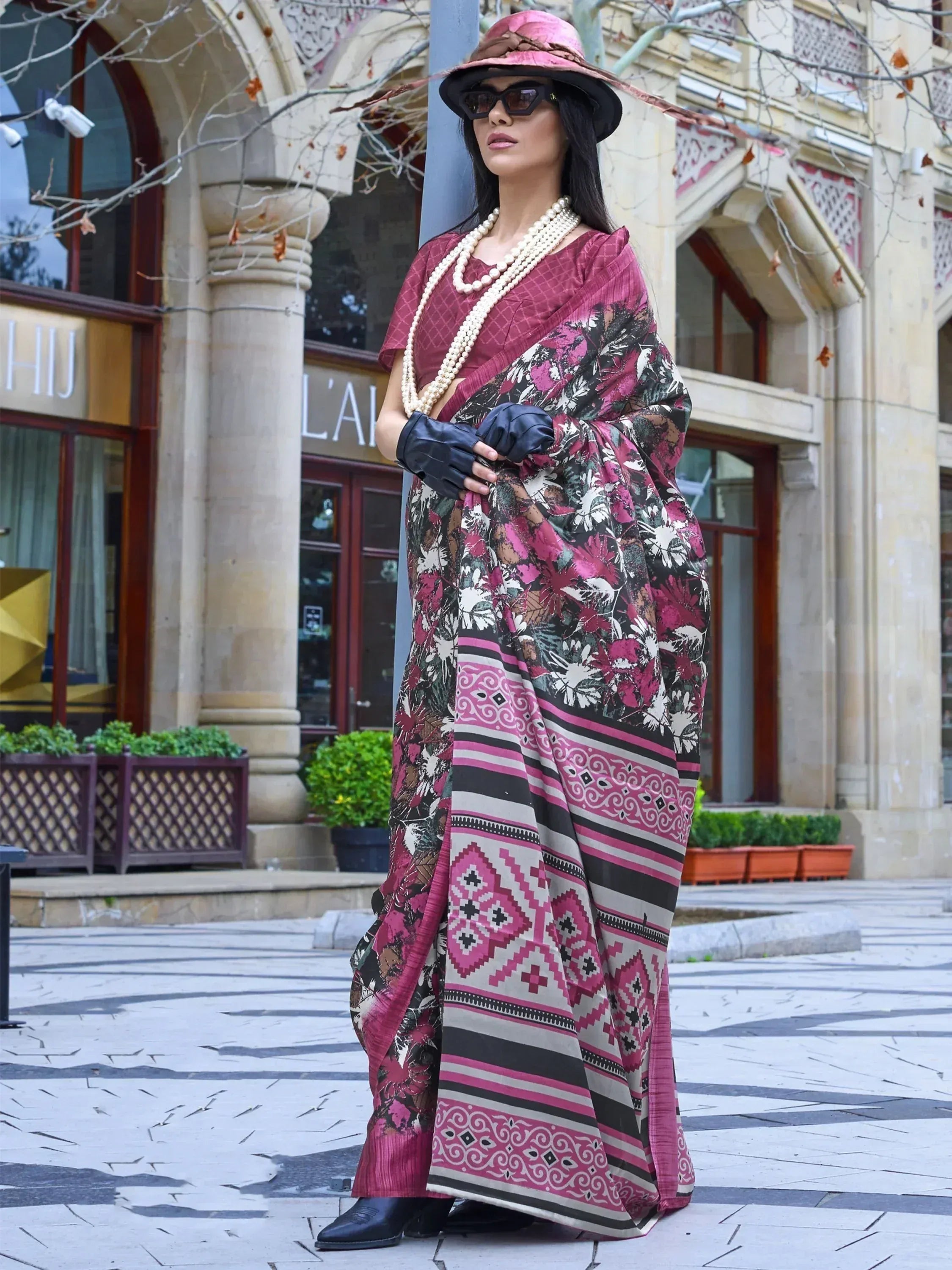 Woman in pink floral printed saree with ethnic patterns, pearl necklace, and wide-brim hat, styled for modern Indian fashion.
