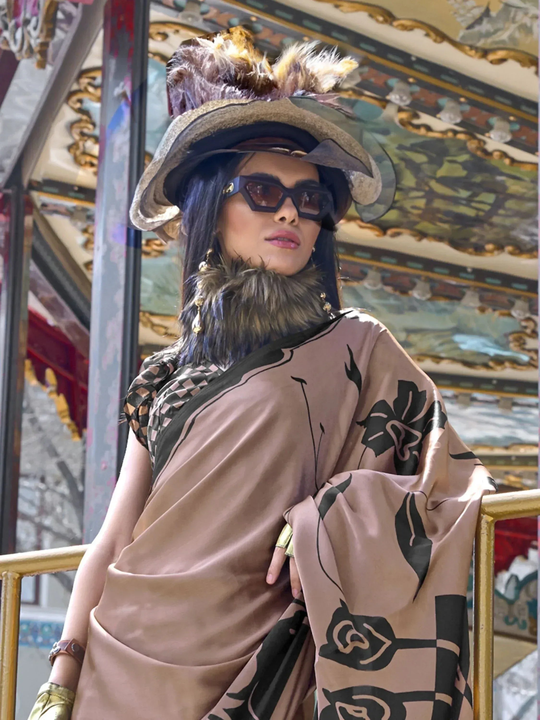 Stylish woman in ethnic gown with floral print, feathered hat, and sunglasses at ornate carousel