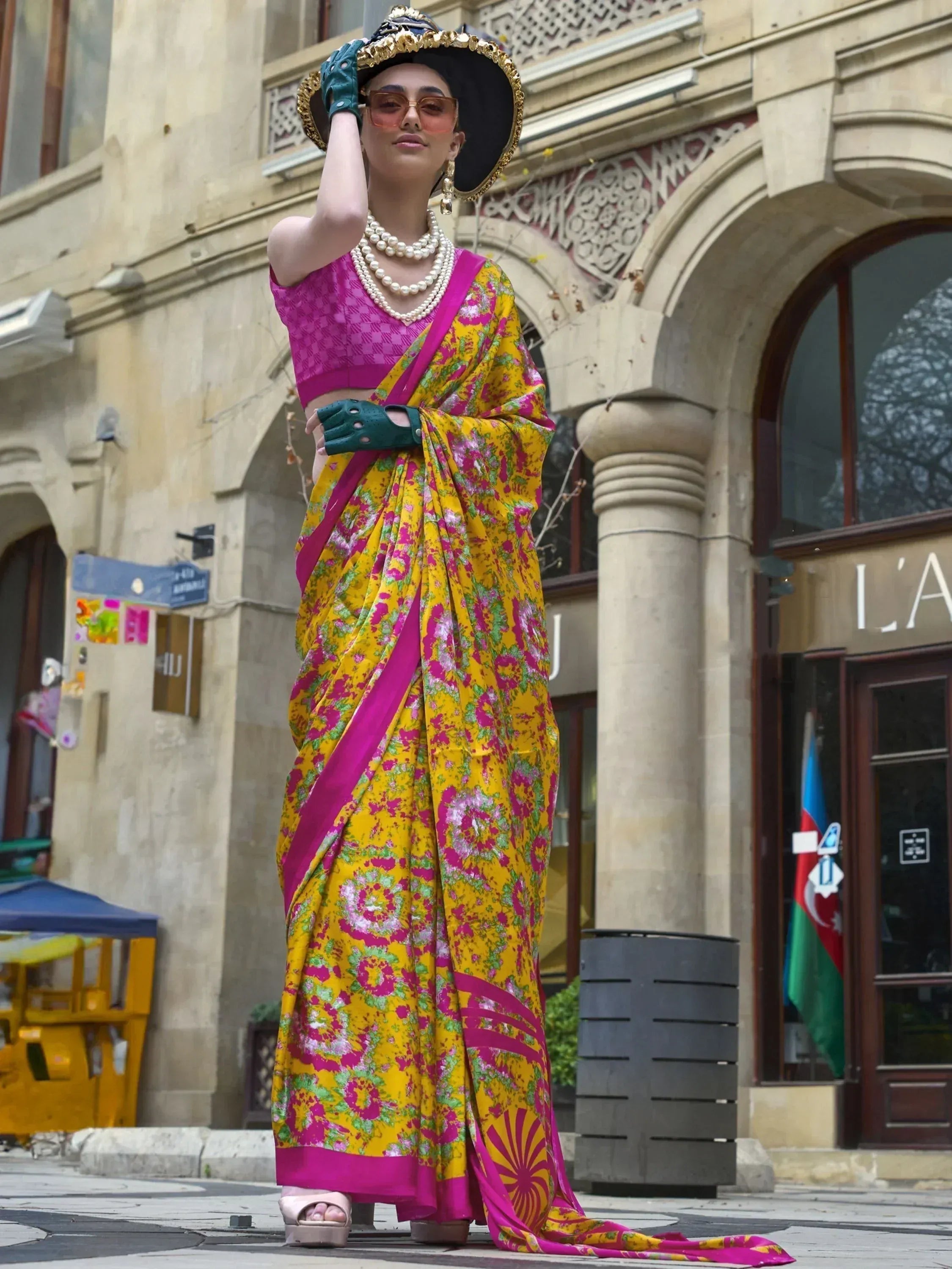 Vibrant yellow and pink floral saree with matching blouse, styled with pearl necklace and gold-trimmed hat for ethnic fashion.