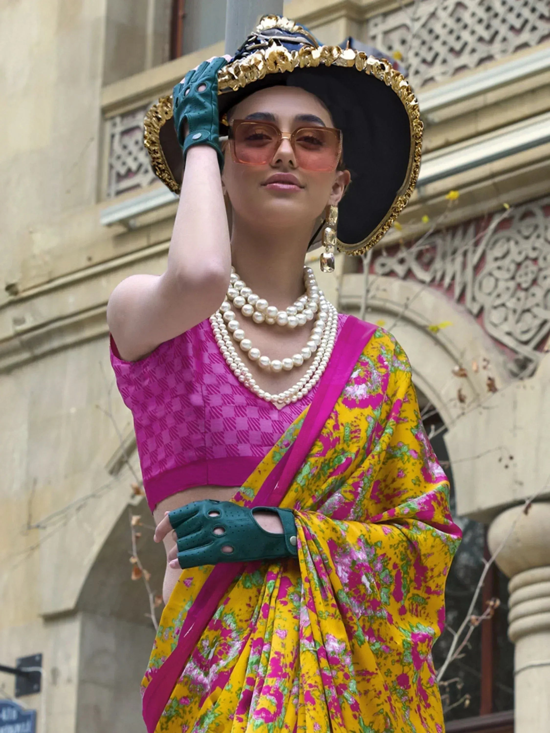 Woman in vibrant yellow floral saree with pink blouse, pearl necklaces, and gold-trimmed hat, styled for ethnic fashion.
