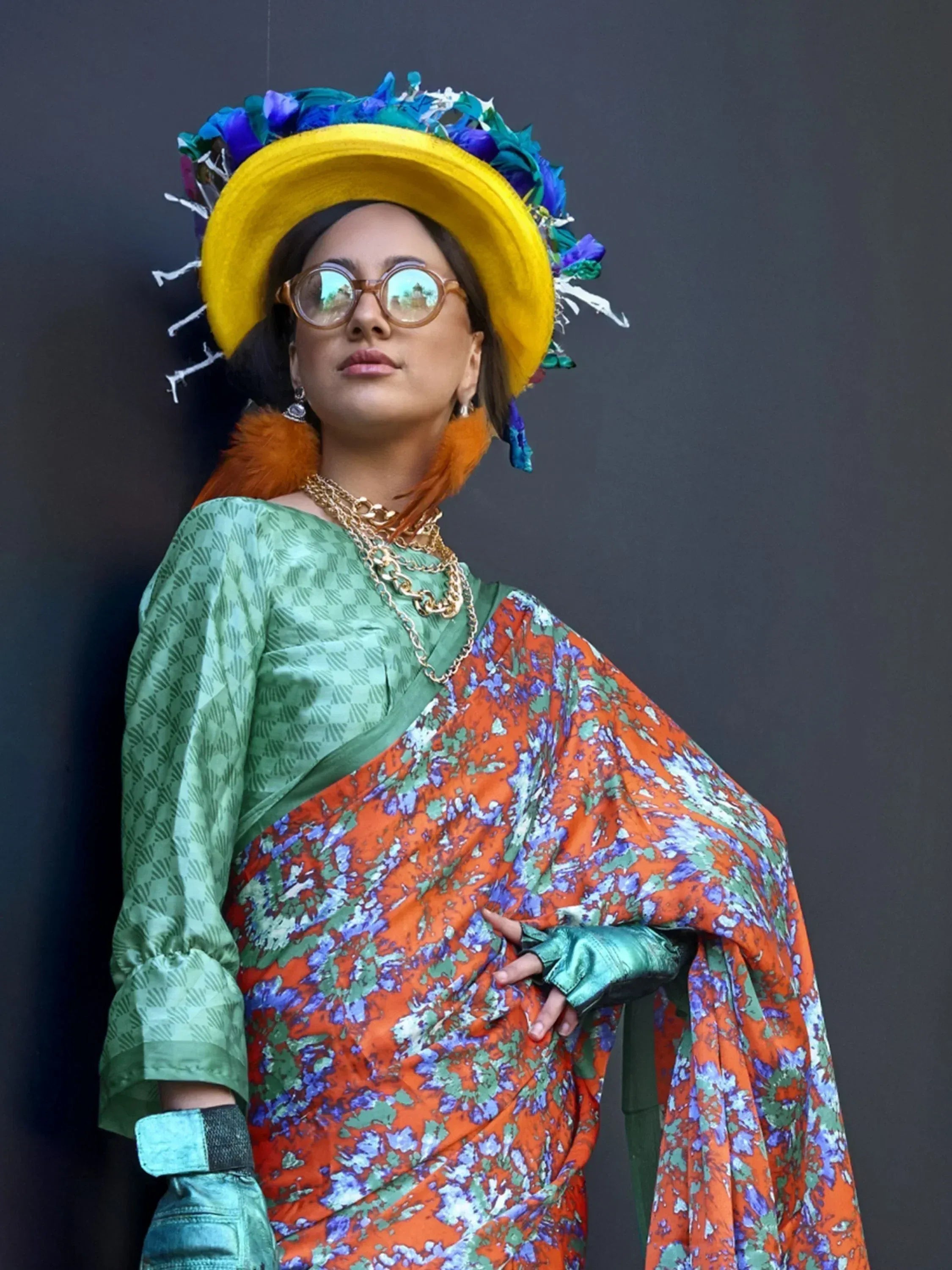 Woman in vibrant floral saree with green blouse, yellow feather hat, and gold jewelry, styled for ethnic fashion.