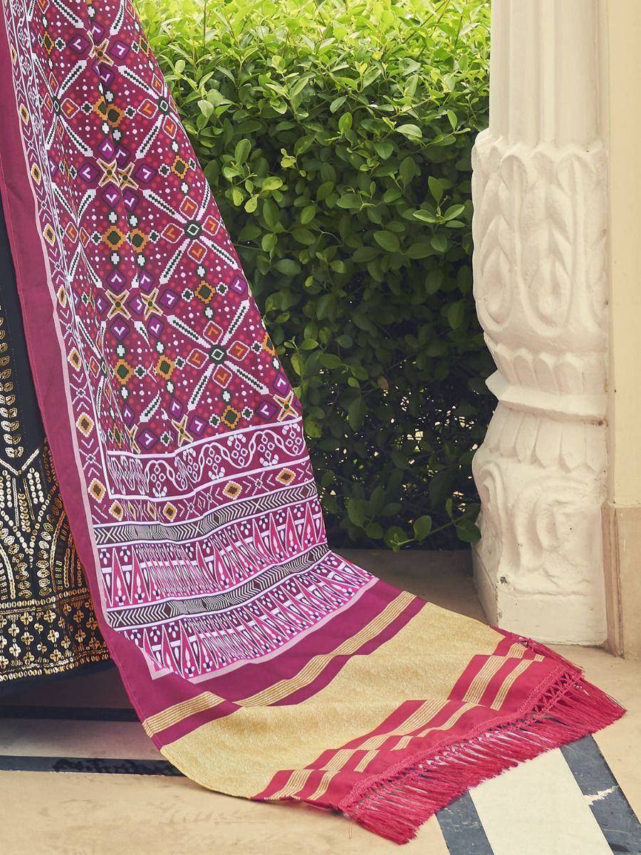 Vibrant maroon ethnic dupatta with geometric prints, gold stripes, and fringe, styled near carved pillar and greenery