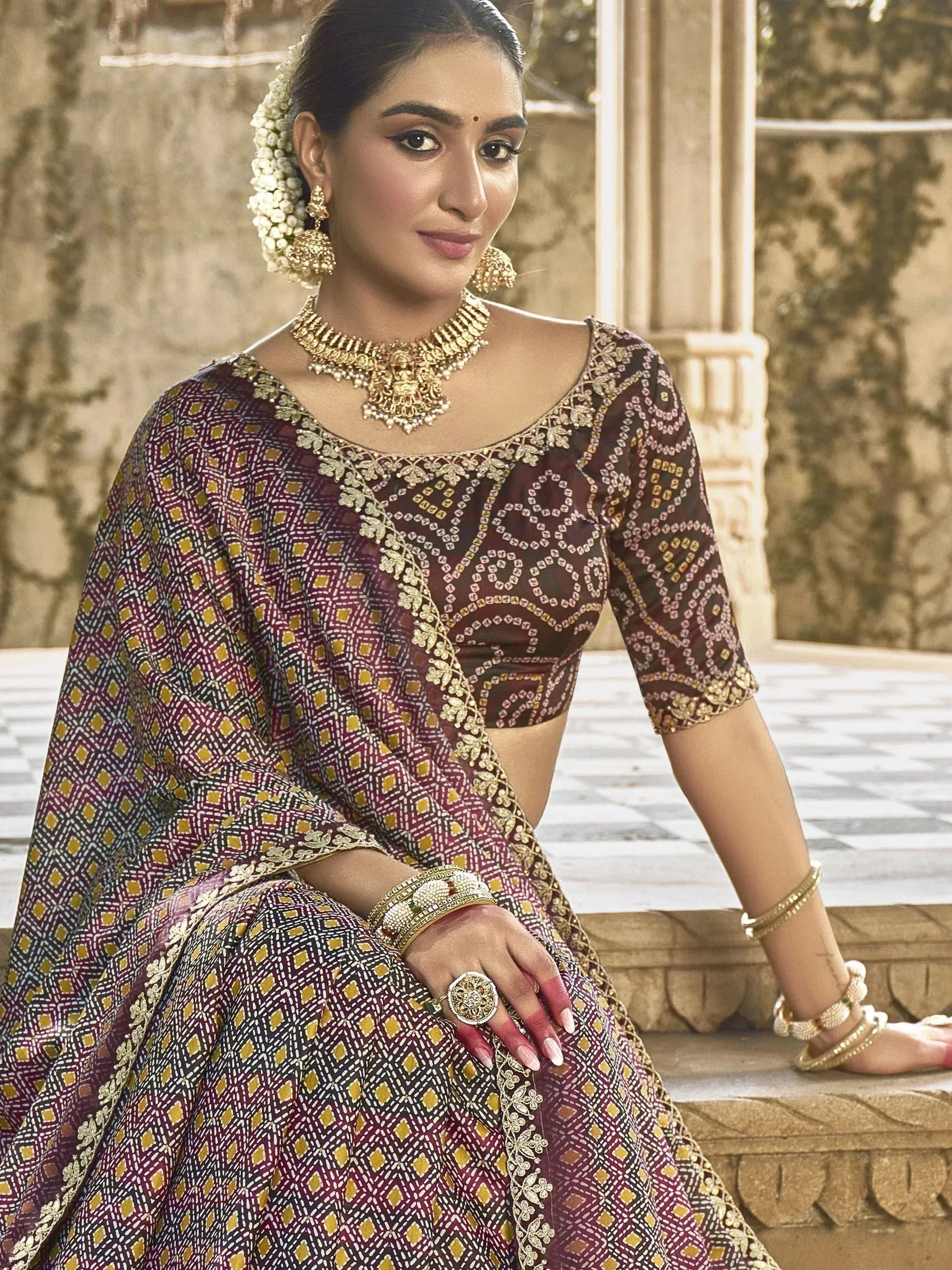 Woman in maroon and gold patterned lehenga choli with intricate embroidery, traditional jewelry, and floral hair accessory