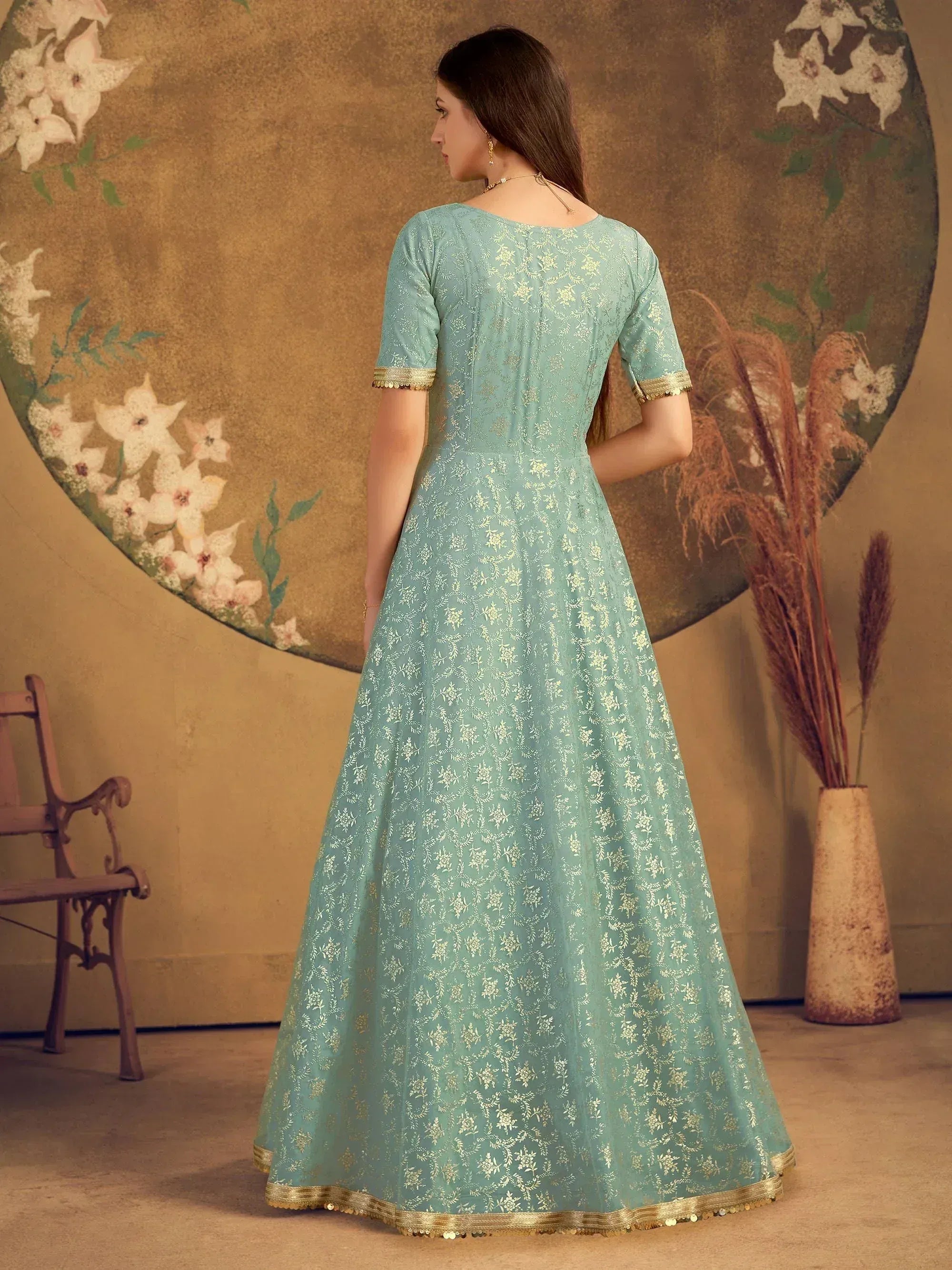 Woman in a light blue floral embroidered gown with gold trim, standing indoors