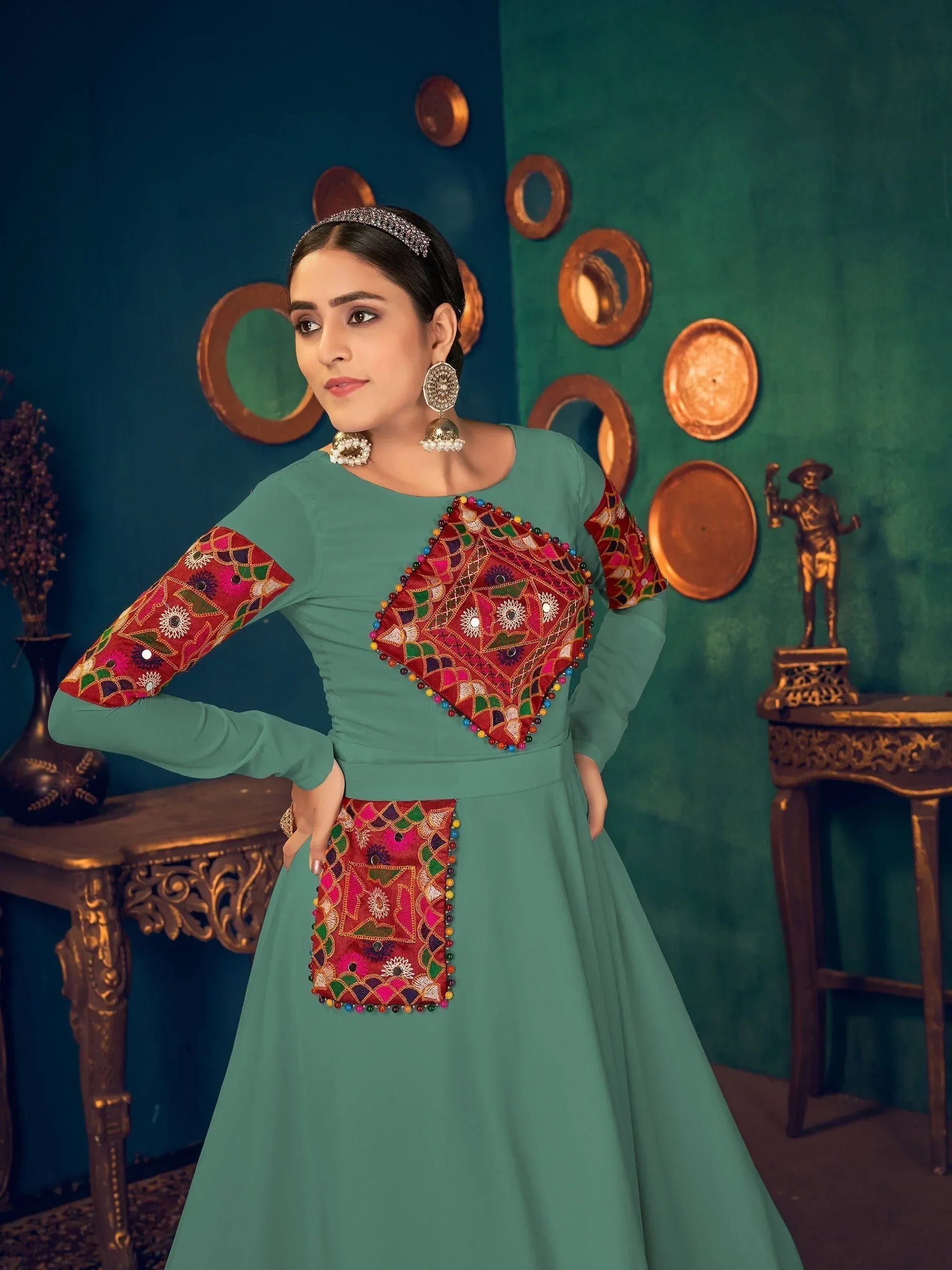 Woman wearing teal ethnic gown with colorful embroidered patterns, statement earrings, elegant décor backdrop