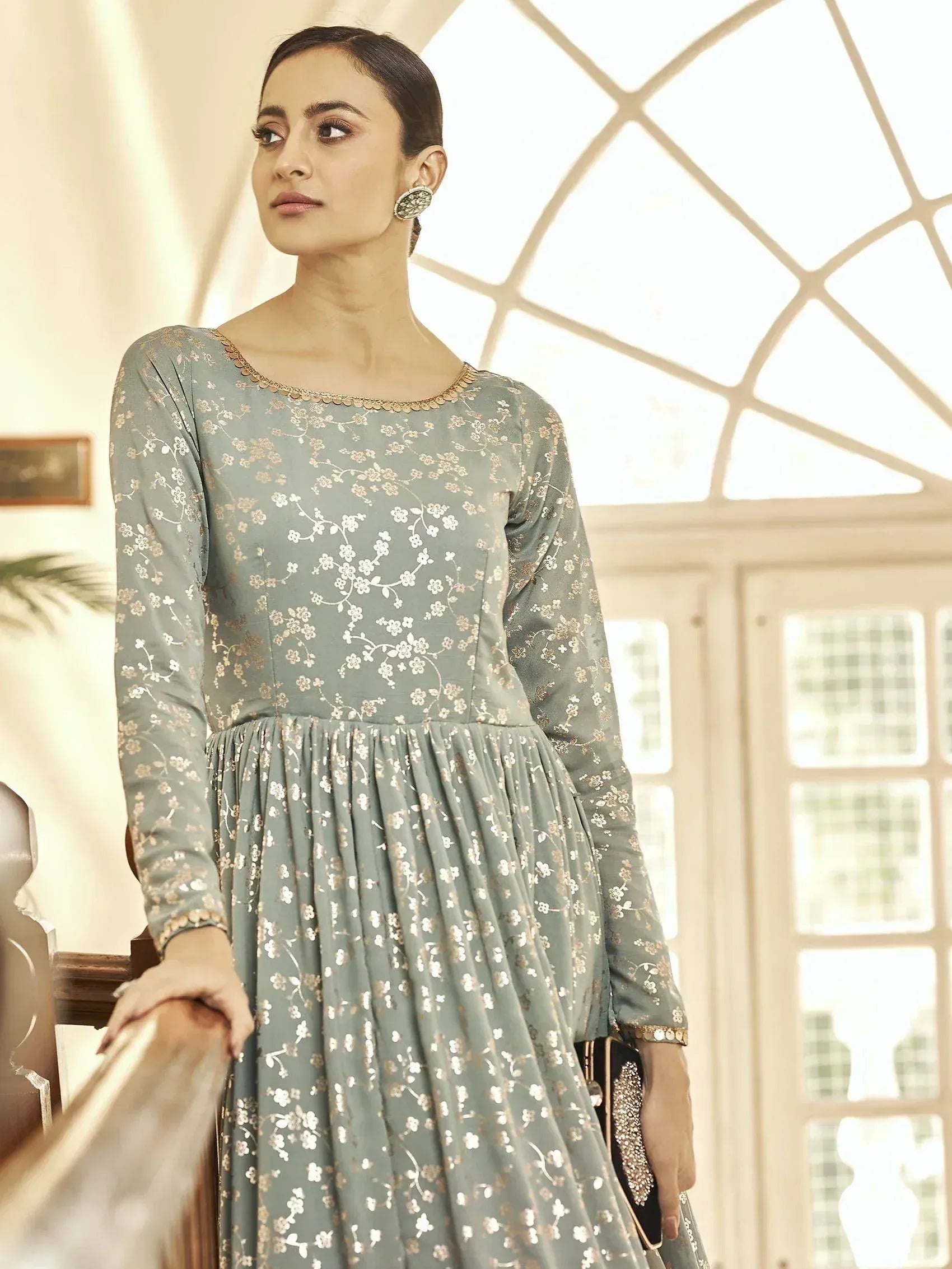 Woman in pastel blue floral print long ethnic gown with full sleeves, elegant earrings, and a clutch.