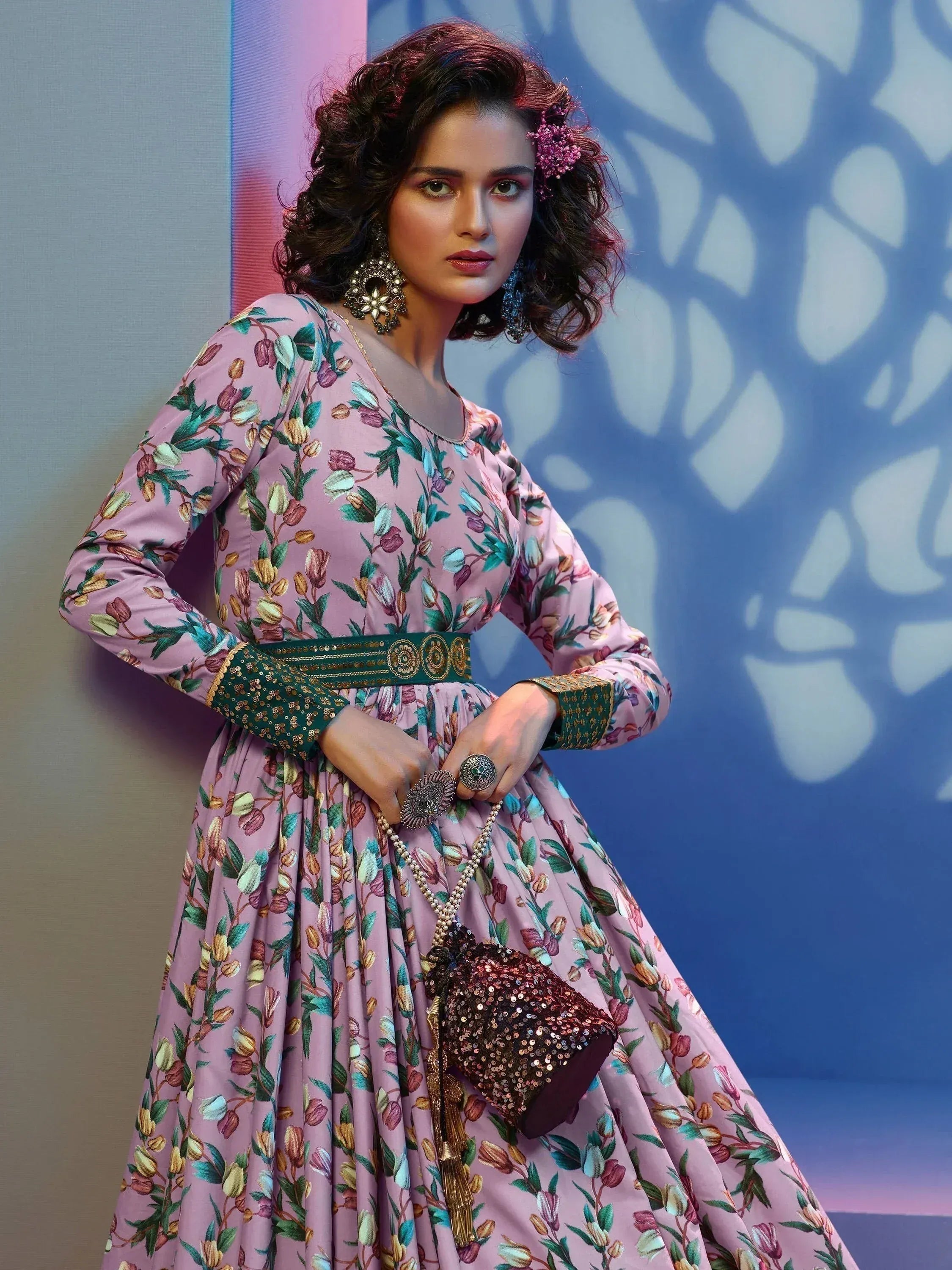 Pink floral ethnic gown with green belt, statement jewelry, and sequin clutch for festive occasions