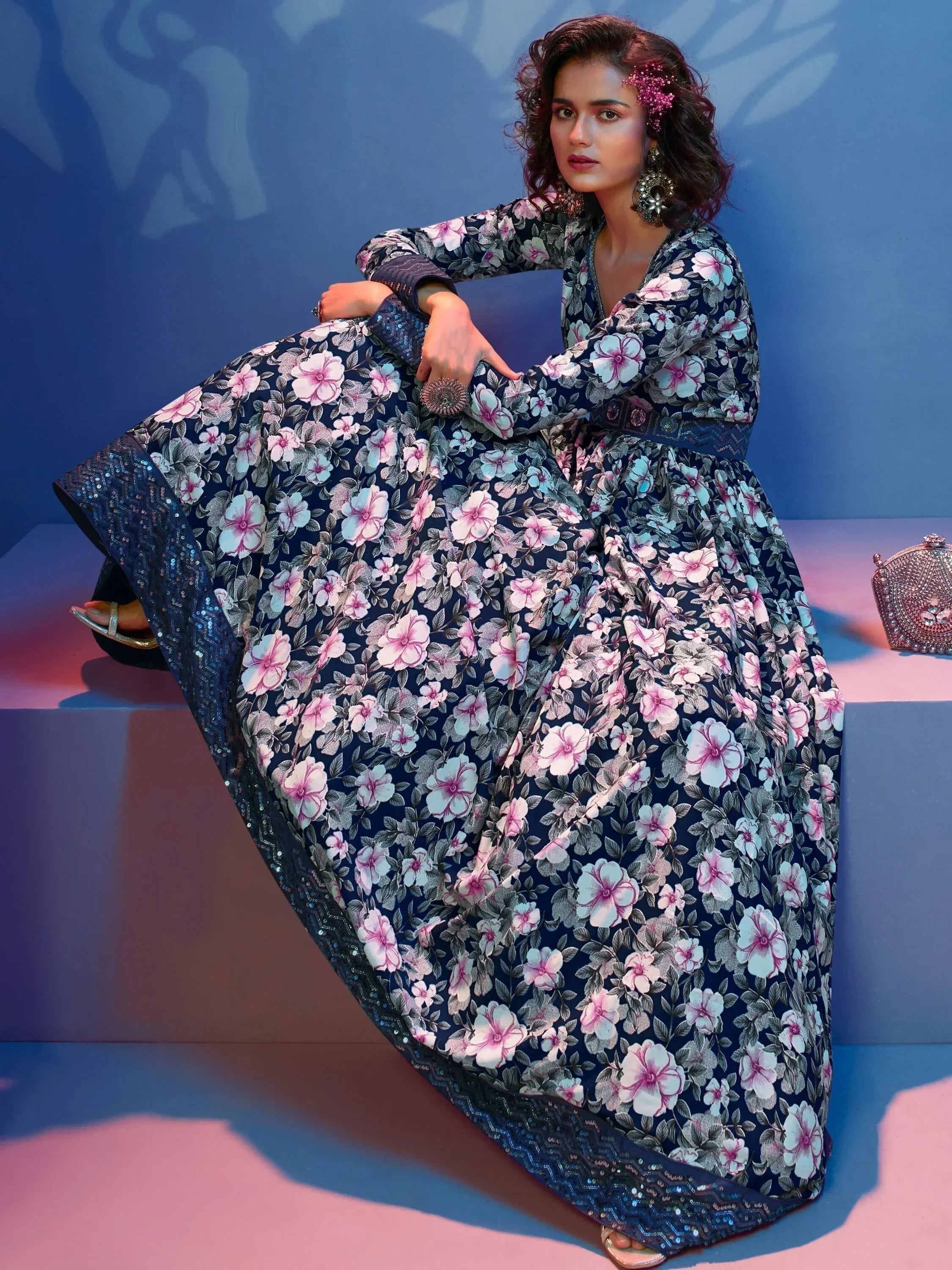 Woman in a navy blue floral print lehenga gown with statement jewelry, seated against a blue background.