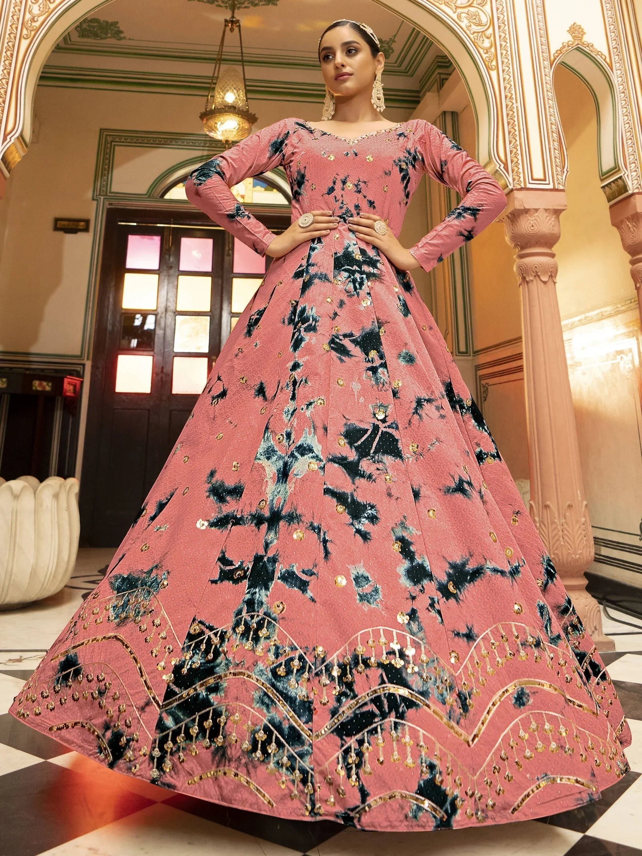 Woman in a pink and blue embroidered designer gown with gold accents, standing in a traditional Indian interior