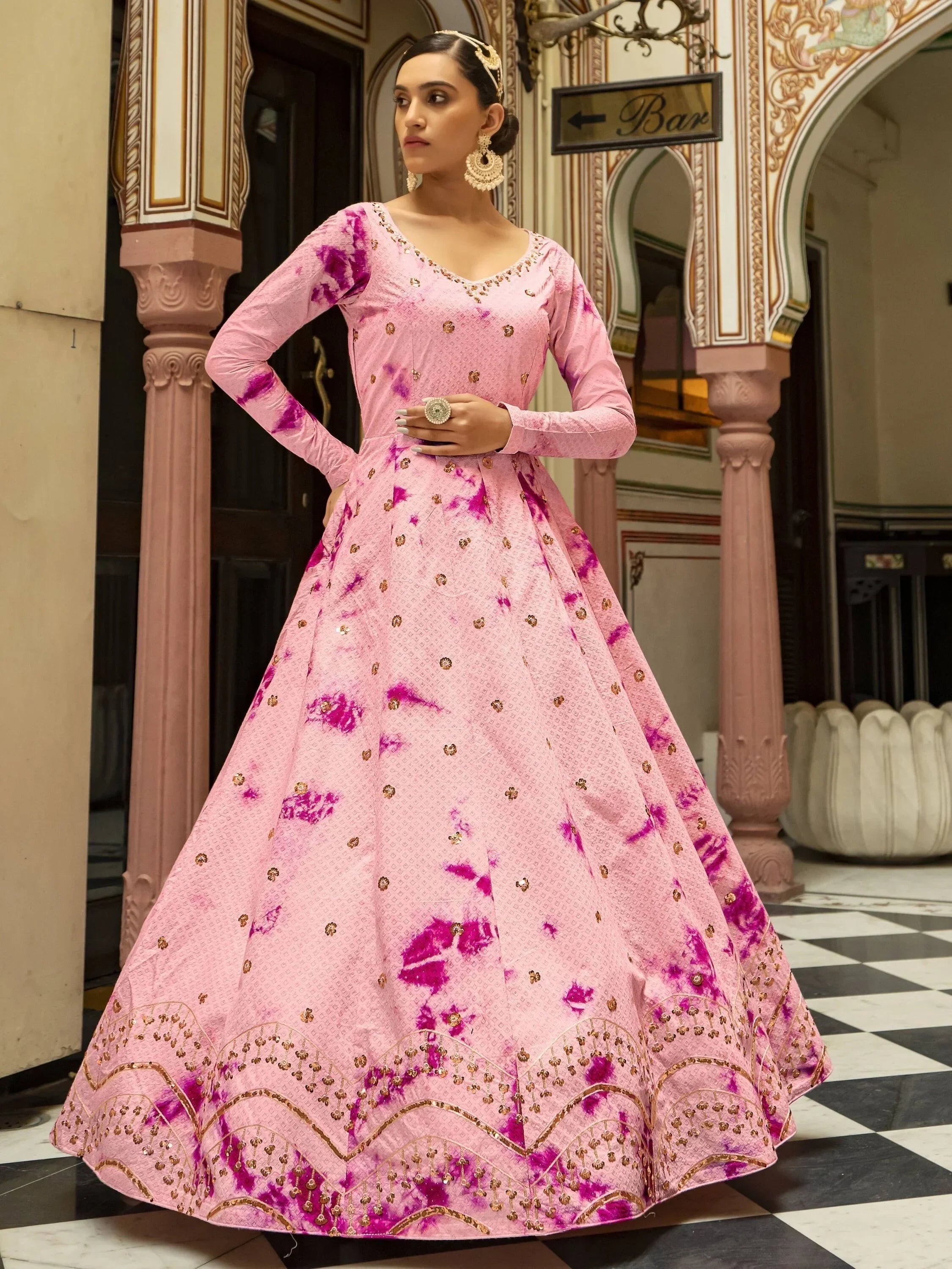 Woman in a pink and purple printed ethnic gown with gold embroidery, traditional earrings, and ring, standing in a heritage-style setting