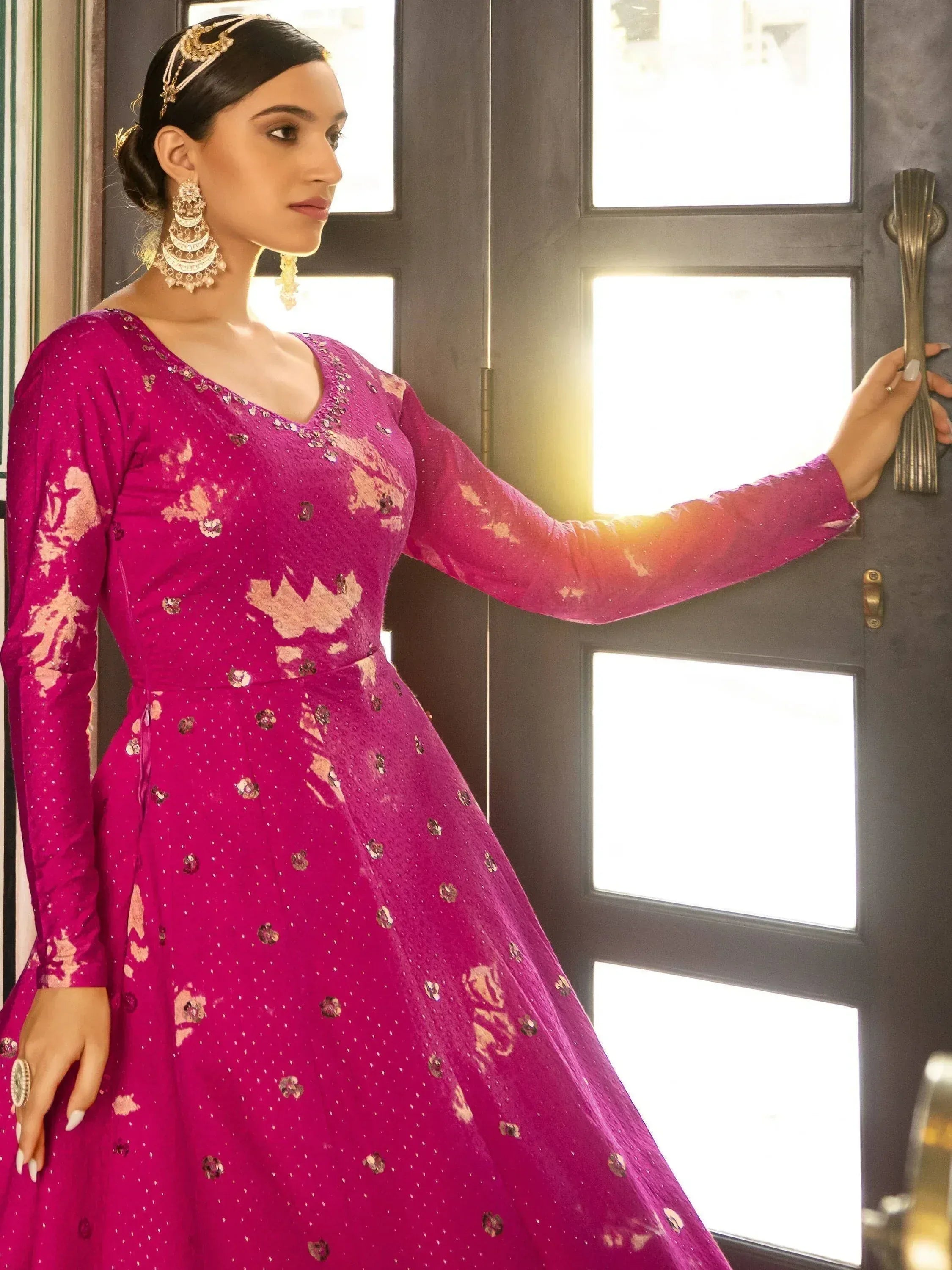 Woman in vibrant pink ethnic gown with gold embroidery, traditional jewelry, and floral motifs, perfect for Indian celebrations.