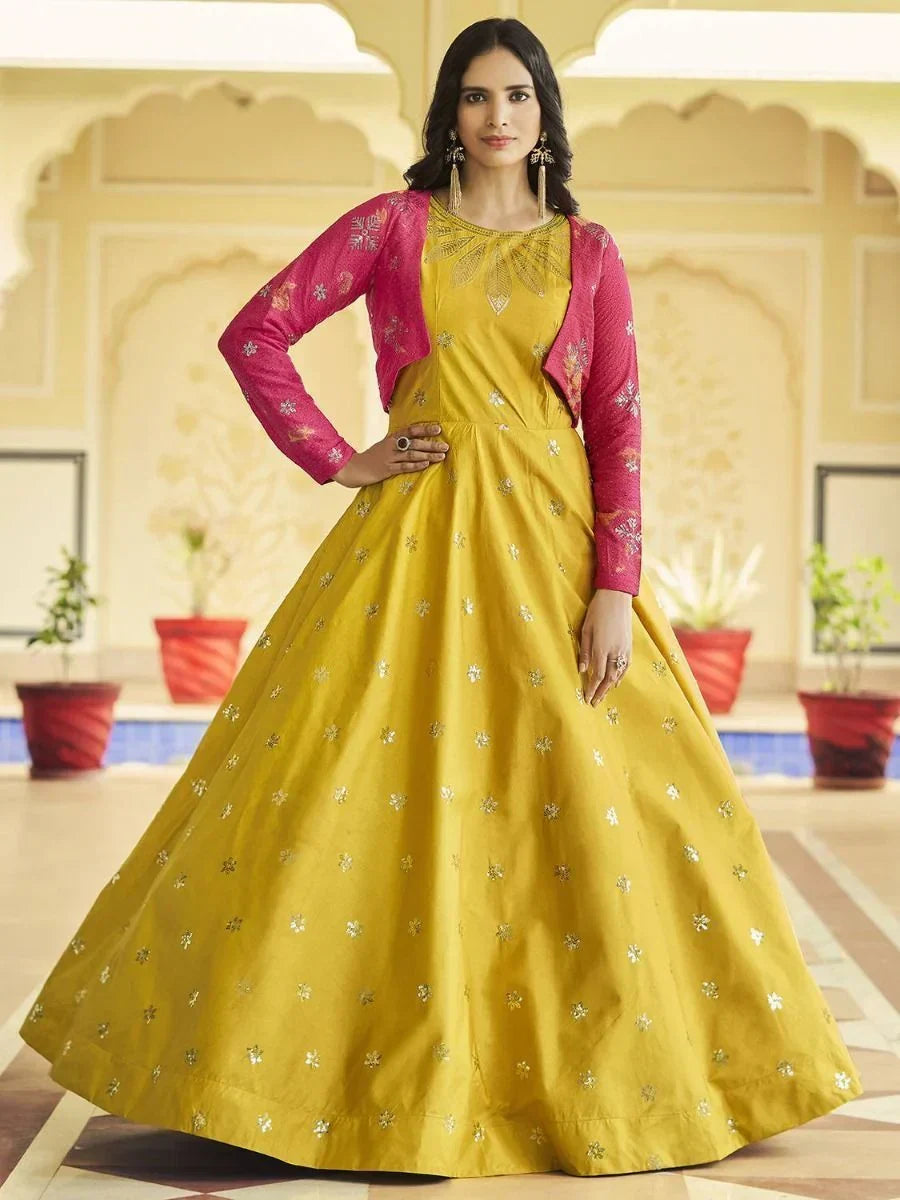 Woman in yellow ethnic gown with silver motifs and pink embroidered jacket, indoor setting