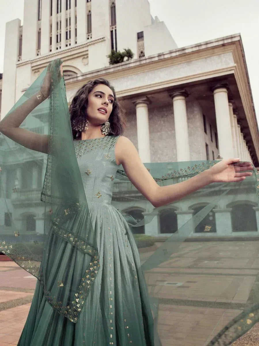 Elegant sage green ethnic gown with gold embroidery, styled with sheer dupatta against heritage architecture