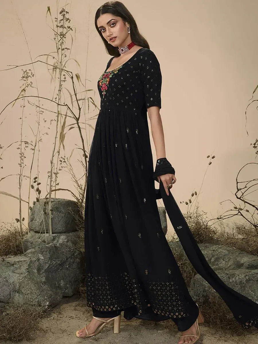Woman in a black Indian ethnic gown with gold embroidery and floral accents, standing outdoors.
