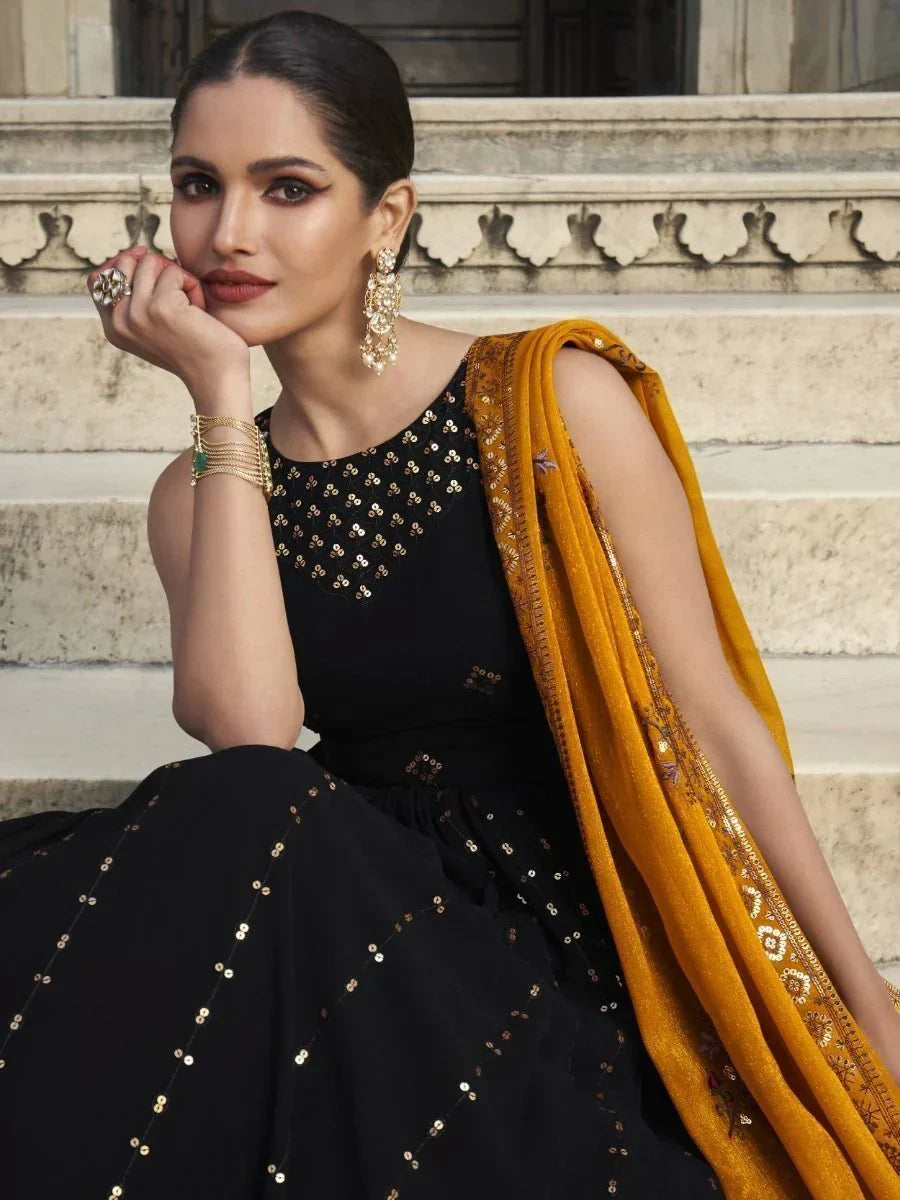 Woman wearing black embroidered ethnic gown with mustard dupatta, traditional jewelry, seated on stone steps