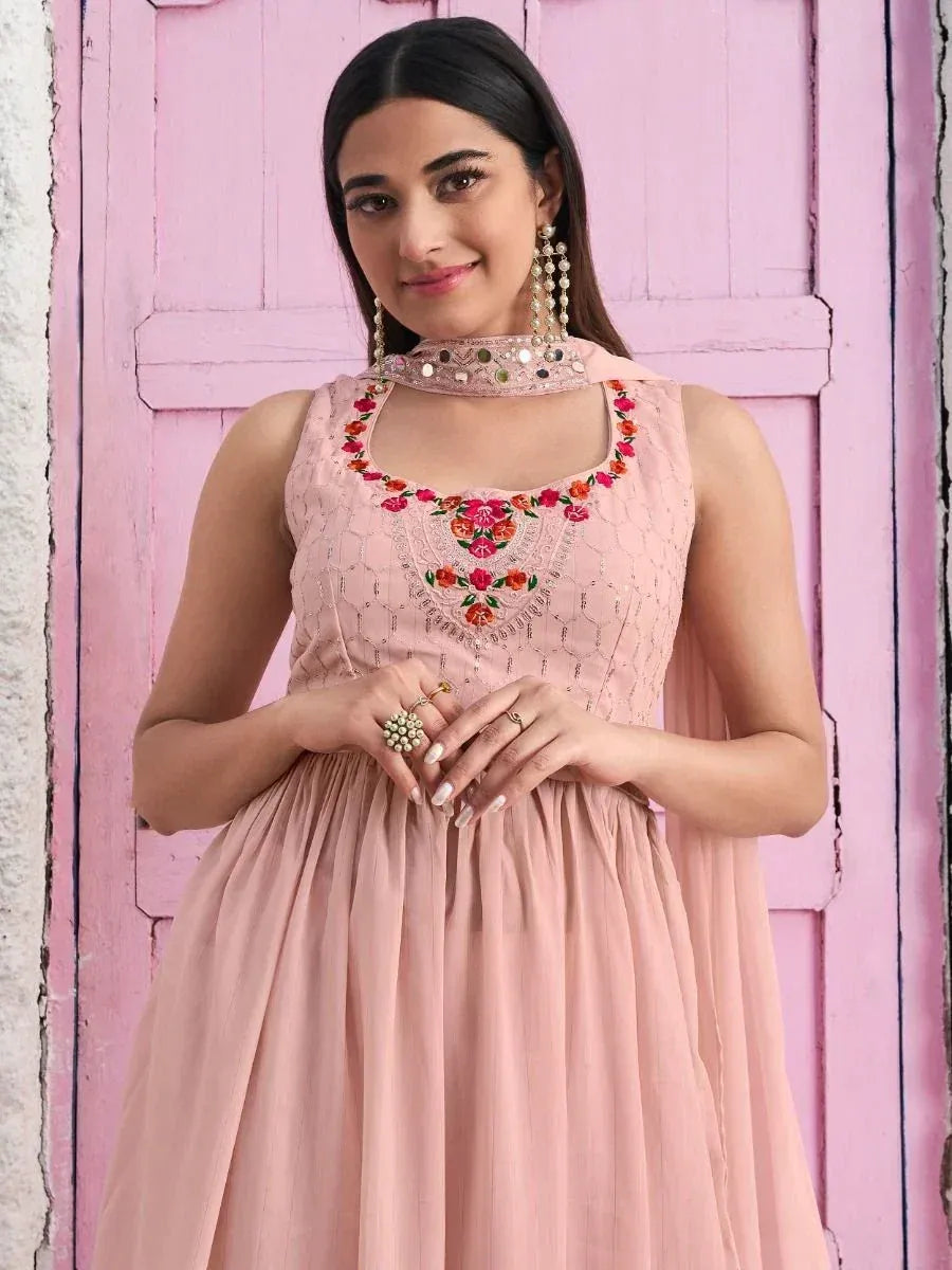 Woman in pink ethnic gown with floral embroidery and dupatta, posing by a pastel door