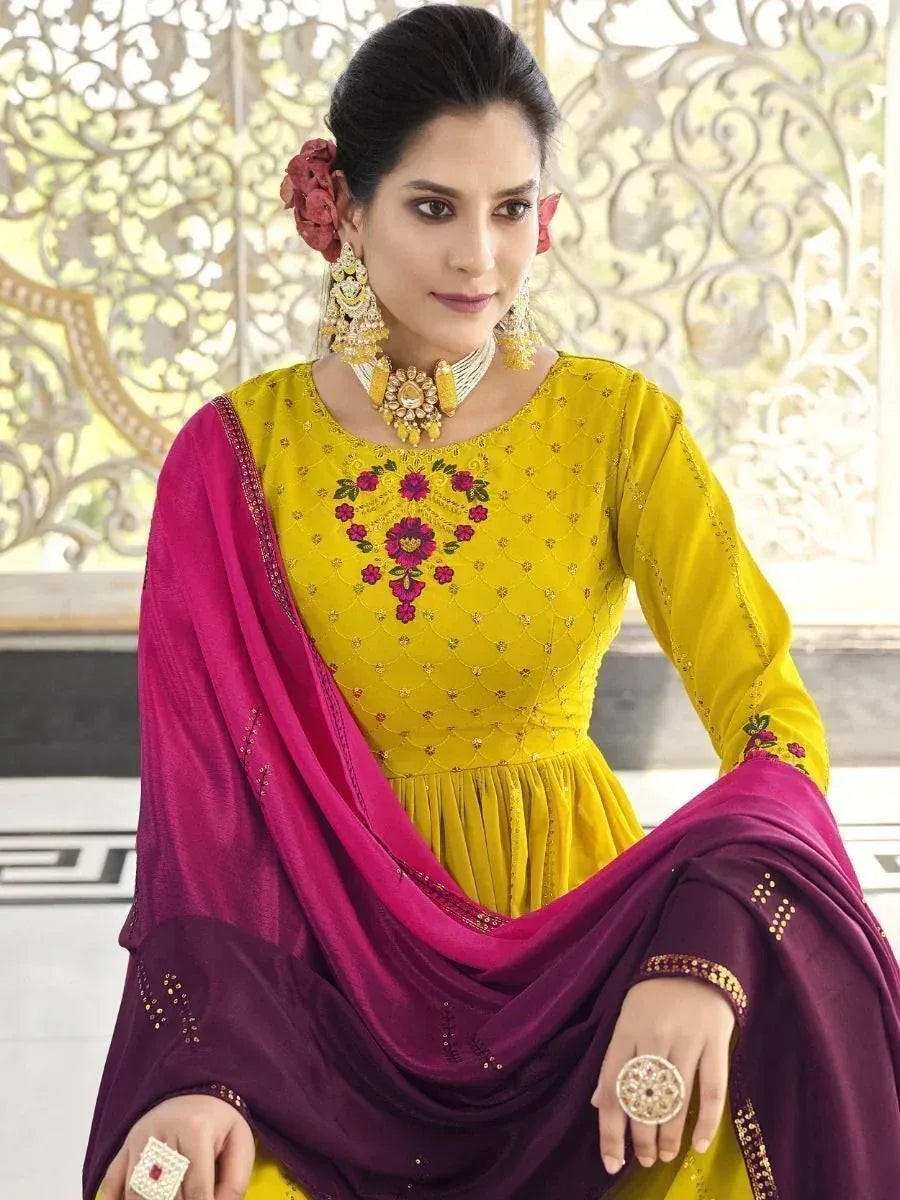 Yellow embroidered ethnic gown with magenta dupatta, floral detailing, and gold jewelry for Indian wear