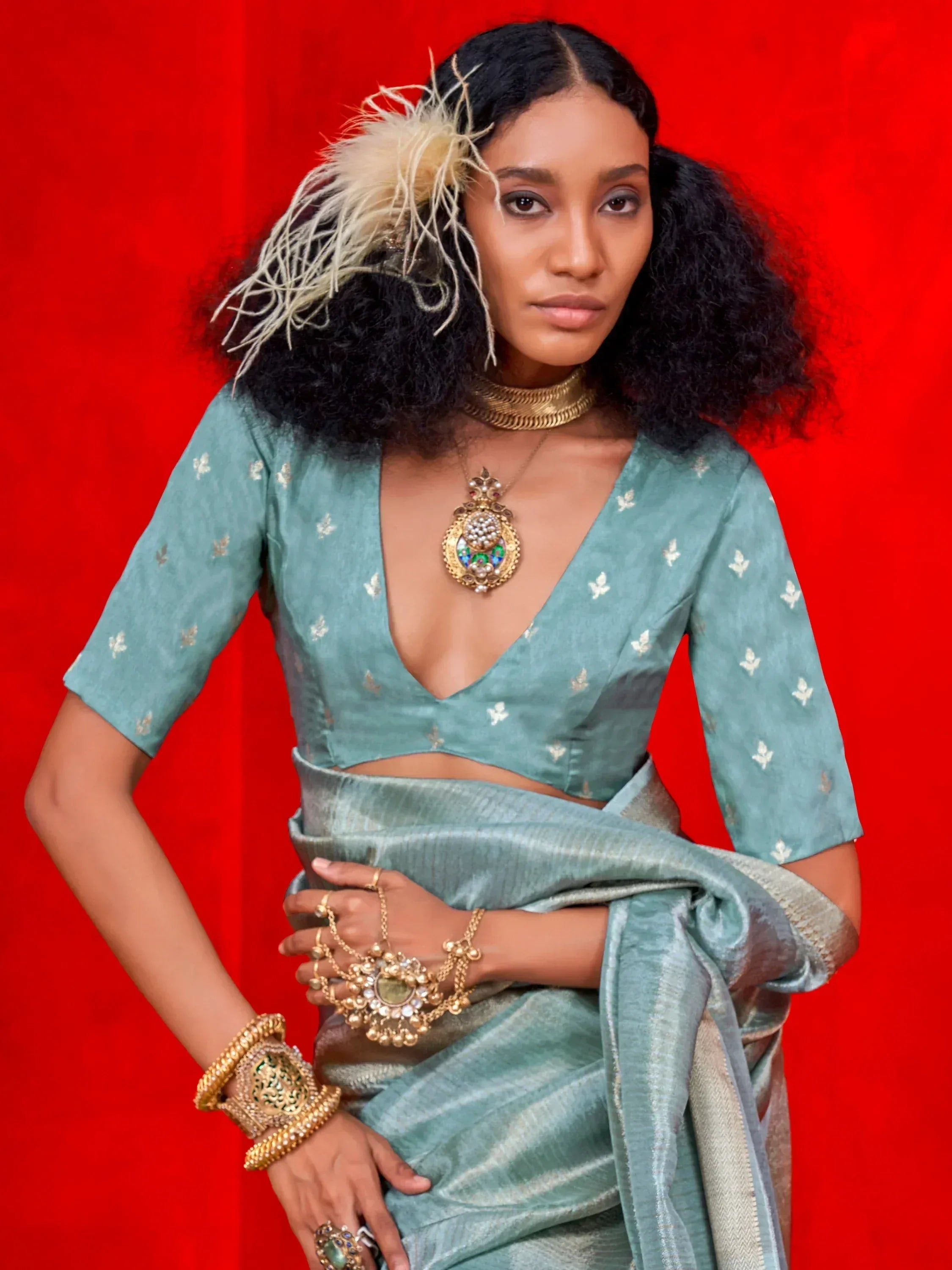 Woman in teal designer saree with gold jewelry and feather hair accessory against red background