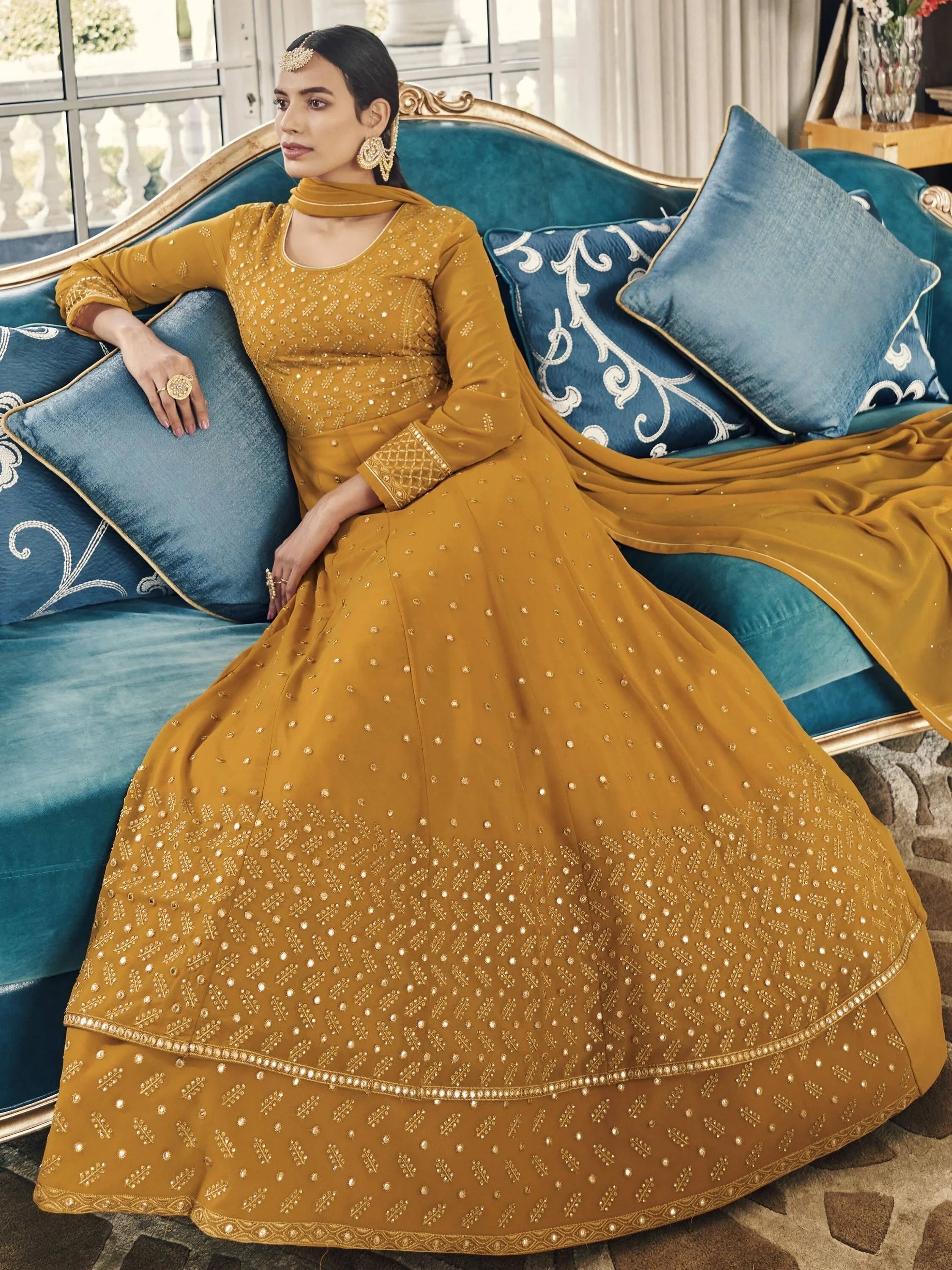 Woman in mustard yellow embroidered anarkali gown with dupatta, sitting on a blue sofa
