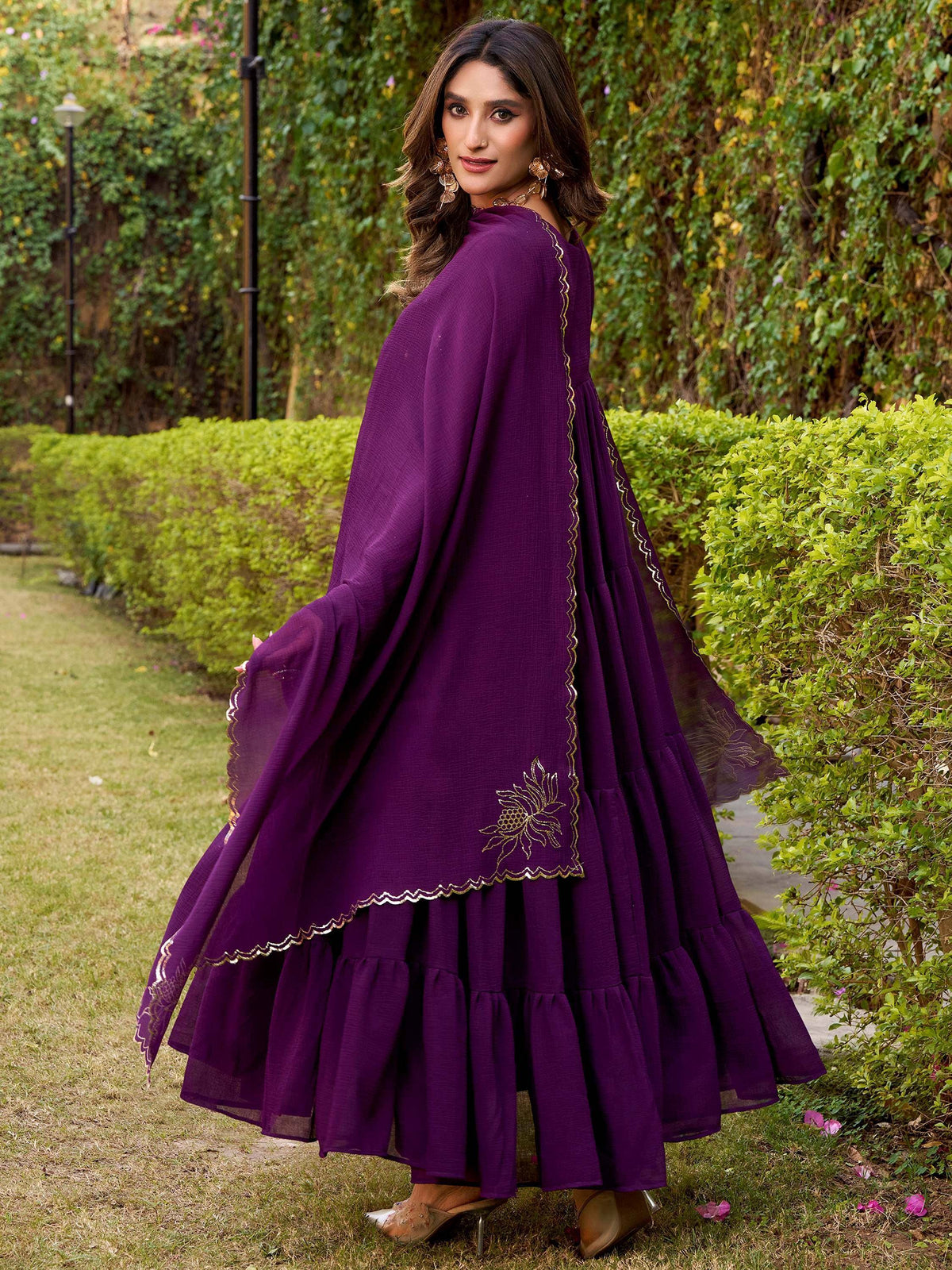 Purple ethnic gown with gold embroidery, flared sleeves, and ruffled layers, perfect for Indian occasions.