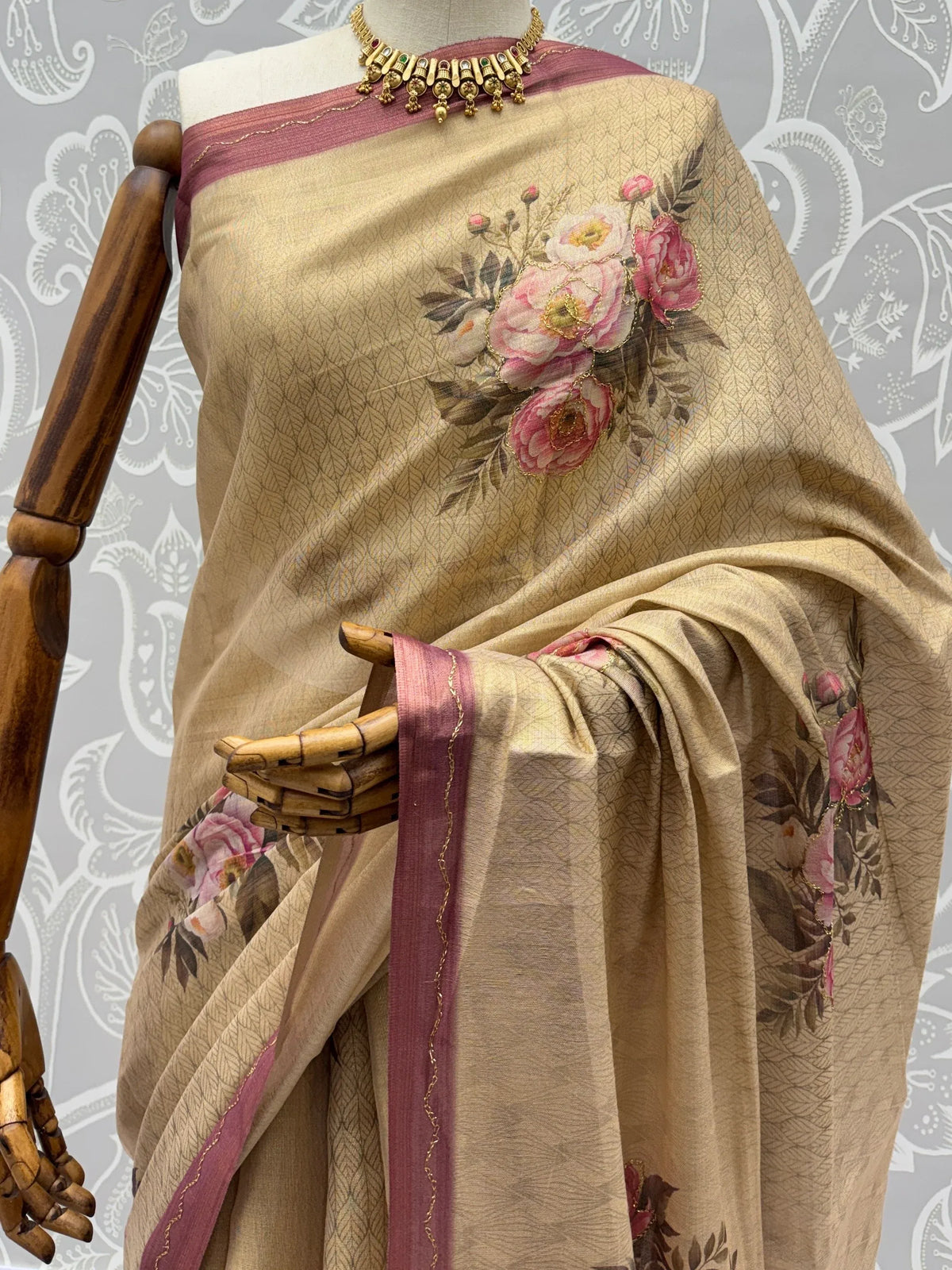 Beige designer saree with pink floral prints and a shimmering border on a mannequin.