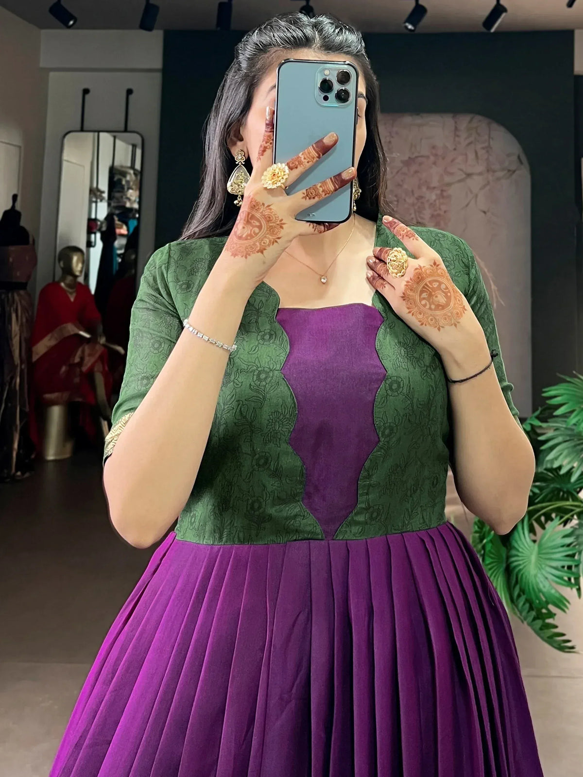 Woman in purple and green ethnic gown with henna hands, taking mirror selfie in boutique