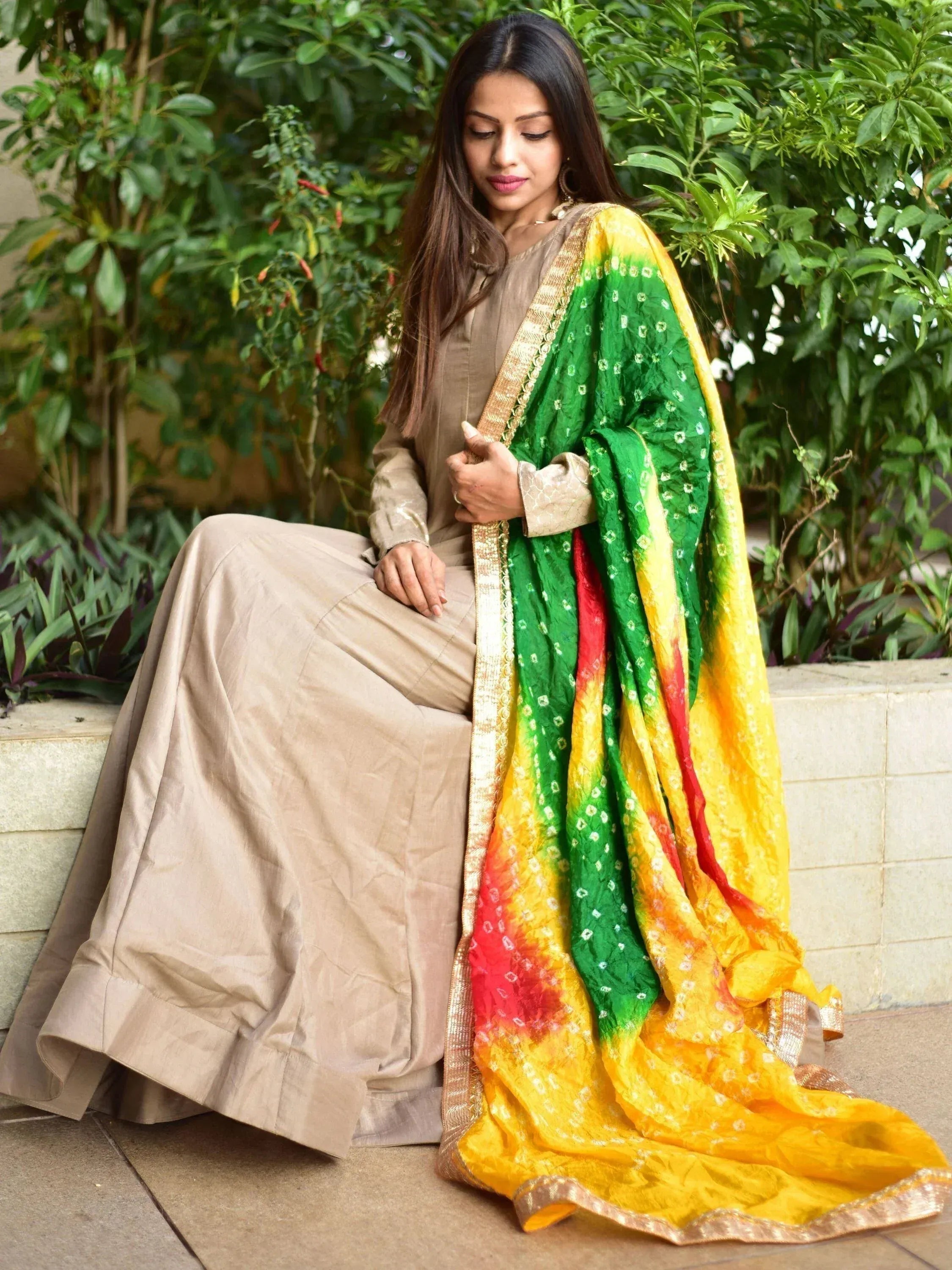Woman in beige salwar kameez with vibrant green, yellow, red tie-dye dupatta, seated outdoors