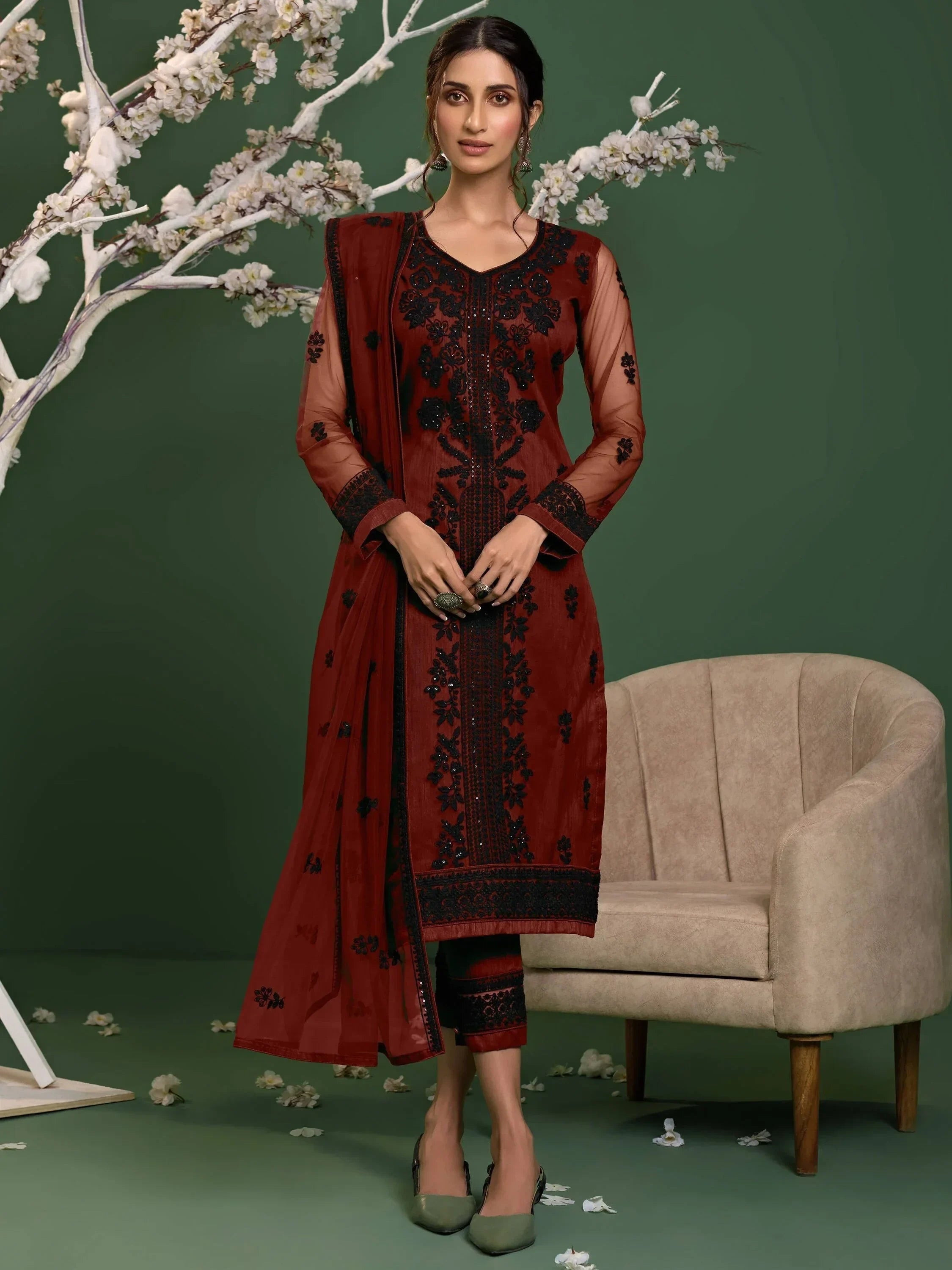 Woman in maroon embroidered salwar kameez standing by a beige chair and flower tree